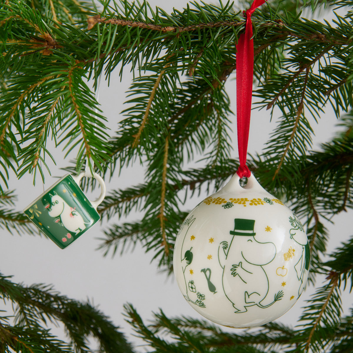 A white ceramic Christmas ornament featuring green and yellow Gift of Love illustrations hangs from a red ribbon on a pine tree branch. A small green mini mug is visible nearby against a soft gray background.