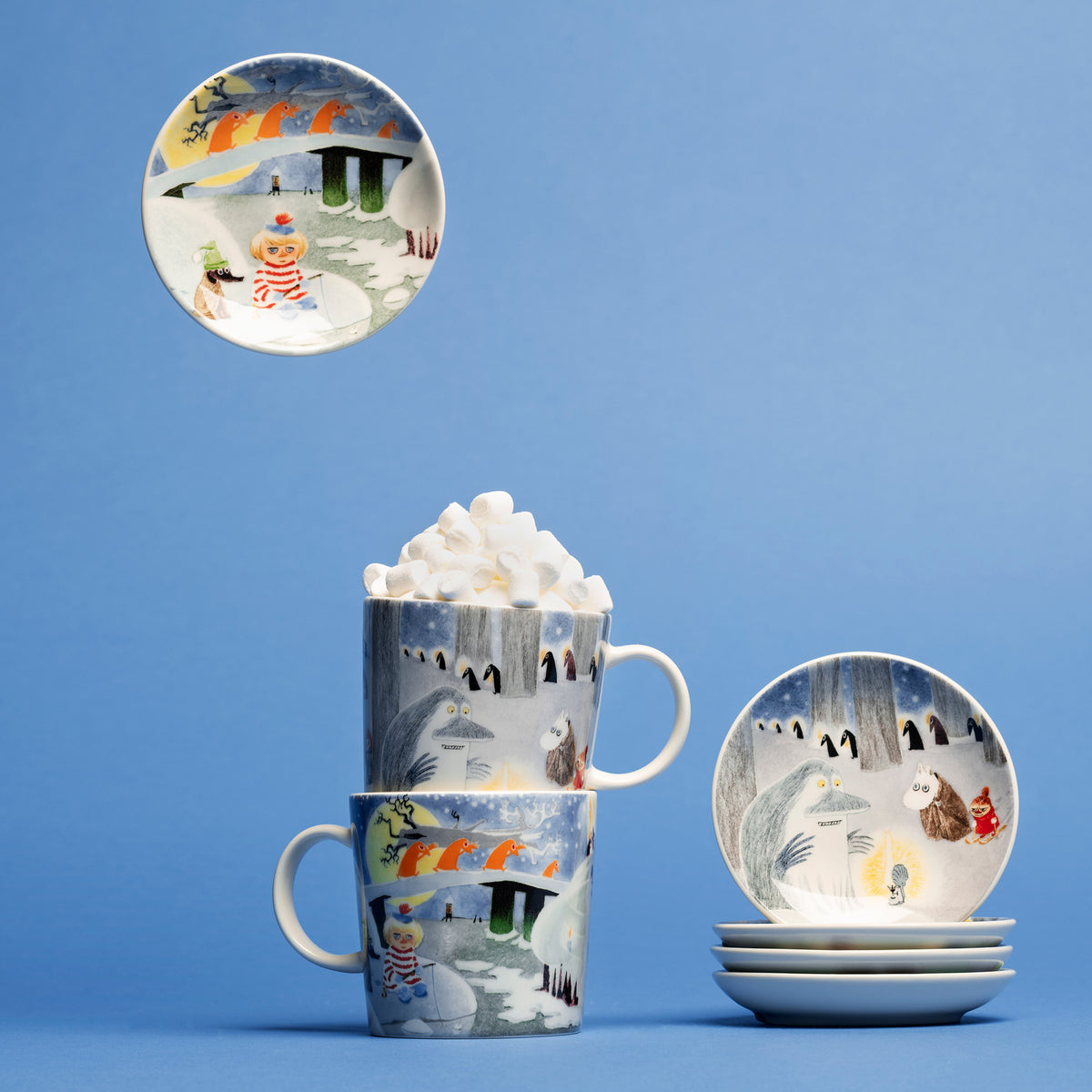 Moomin mini plate 10,5cm Winternight glow 2pc - A set of ceramic tableware featuring a Winternight Glow illustration, displayed on a solid blue background. The set includes mugs topped with whipped cream, a stack of mini plates, and a mini plate showing the same illustration hanging on the wall.