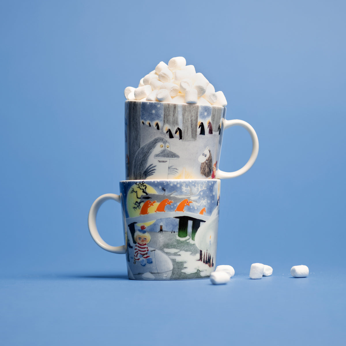Moomin mug 0,3L Winternight glow - Two stacked ceramic Winternight Glow mugs, filled with whipped cream and marshmallows, set against a solid blue background.
