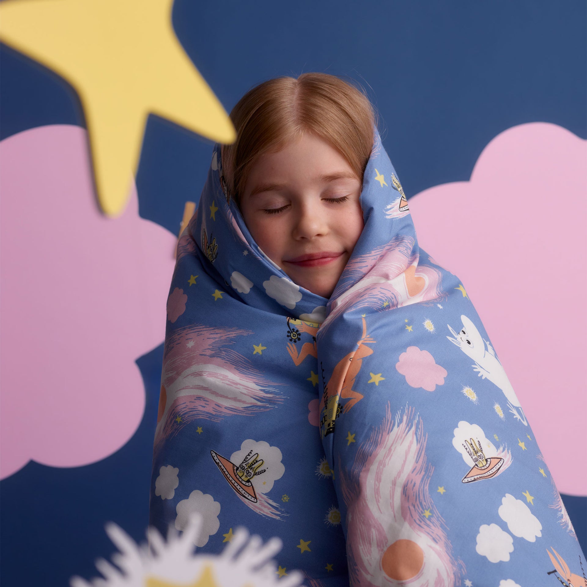Mumin bäddset 120x160cm Rymdäventyr - A child wrapped in a blue Space Adventure blanket, set against a blue and pink cloud backdrop.
