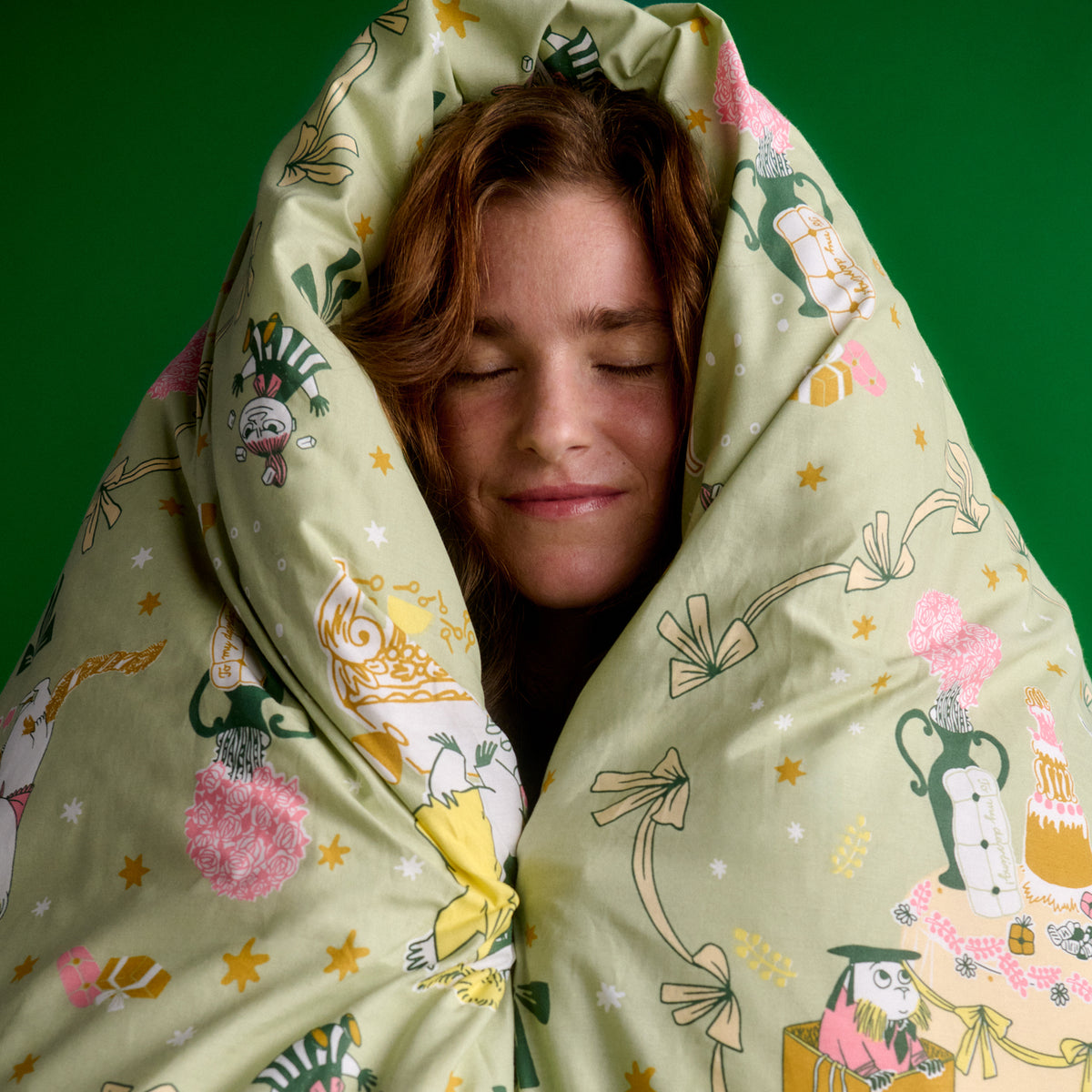 Moomin bedset 140x200cm Gift of Love - A person wrapped in a light sage green patterned duvet or blanket, with their face peeking out against a solid green background. The fabric features Gift of Love illustration.