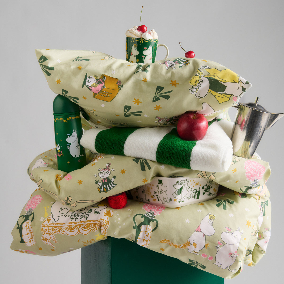 Moomin pillowcase 60x63cm Gift of Love - A stack of Gift of Love bedding and accessories, including a light green patterned duvet cover, matching pillowcases, a green and white striped throw towel, a green mug, and a metal teapot, styled with decorative props like cherries and an apple on a neutral gray background.