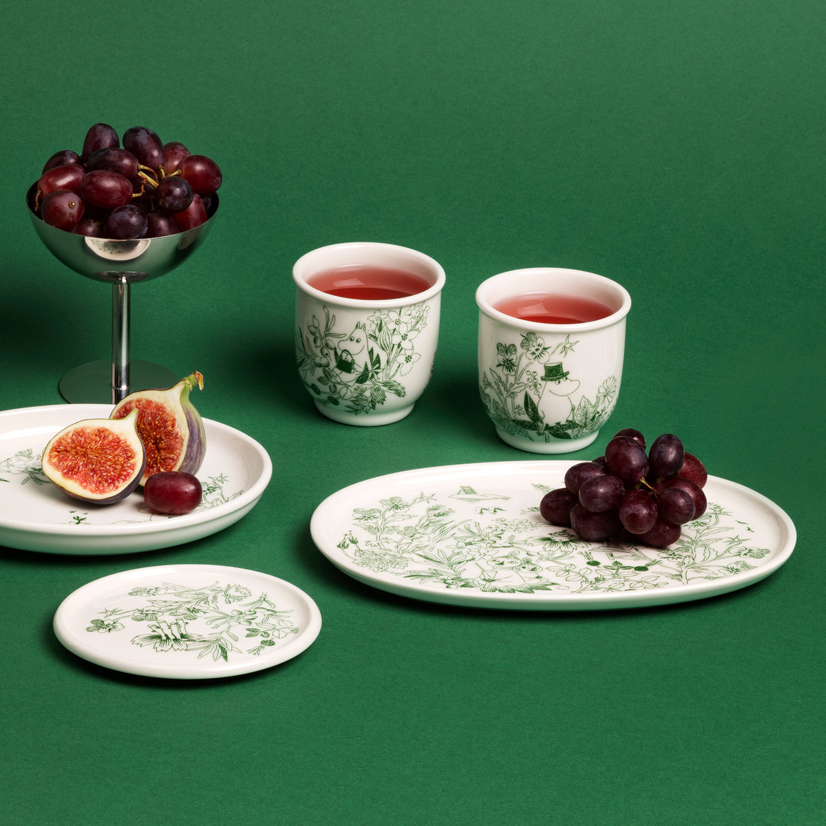 Mumin serveringstallrik 17cm Haru grön - A set of Haru Green ceramic tableware with green botanical illustrations, arranged on a solid green background with fruit props including grapes, figs, and two cups of red drink.