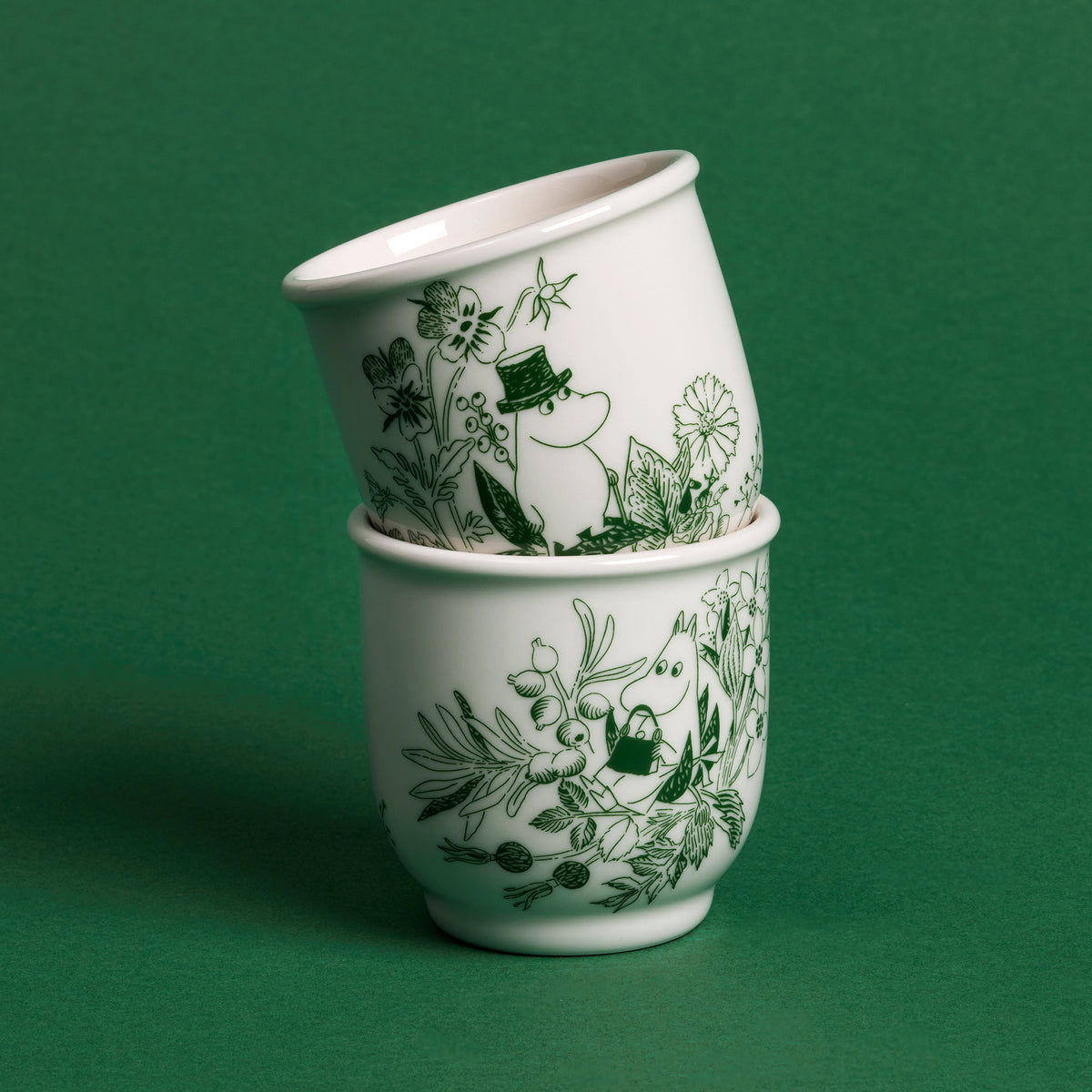 Mumin temugg 26cl Haru grön - Two stacked white tea mugs decorated with a dark green botanical and floral pattern, set against a solid dark green background.