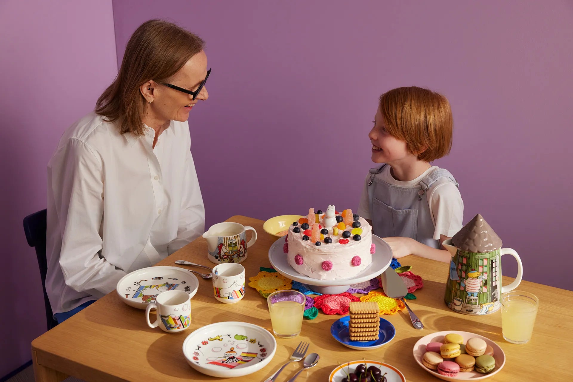 Woman and child at a table with a cake and various Moomin serving items on a purple background