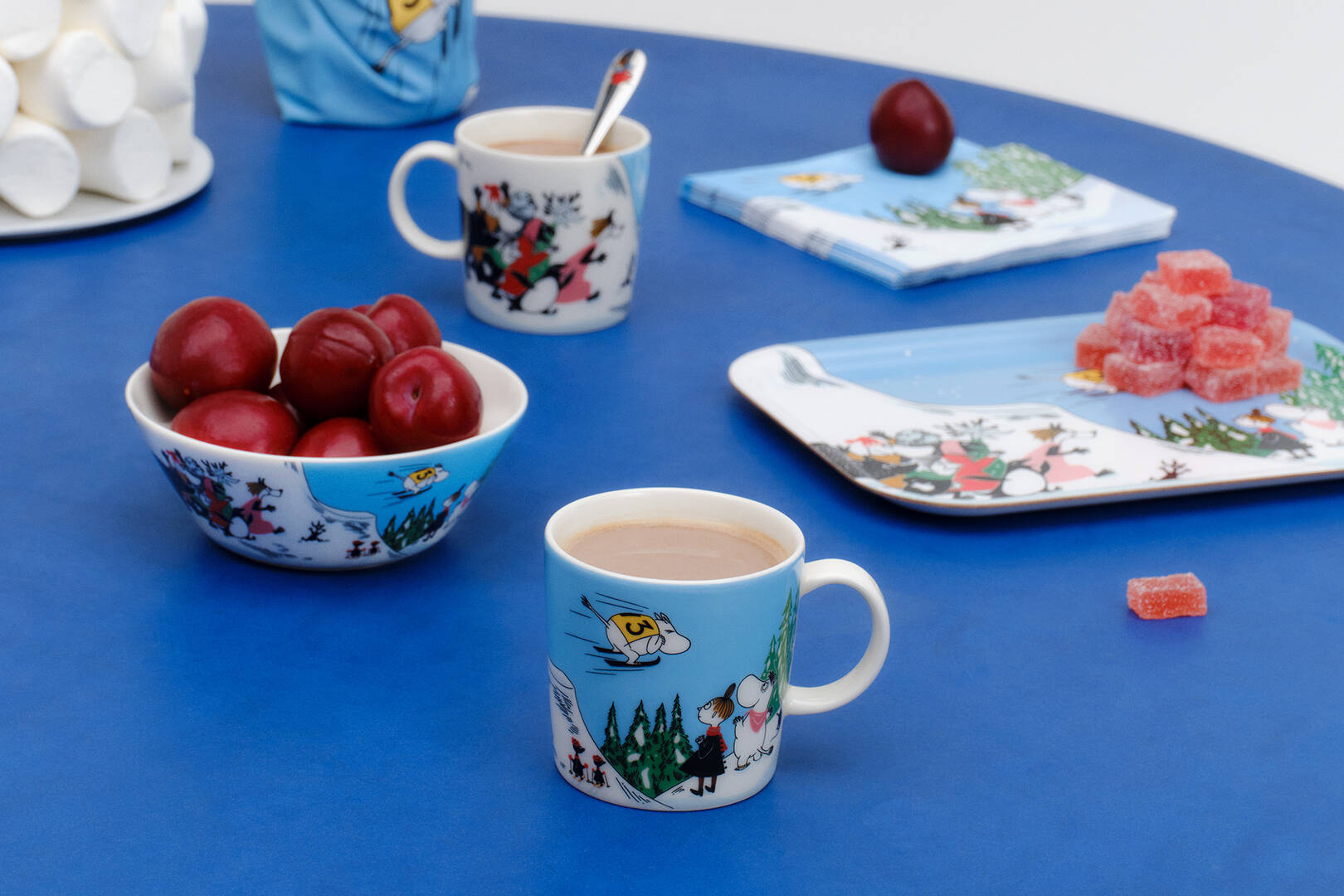 Table setting with Moomin winter seasonal mugs, bowls, and snacks on a blue surface