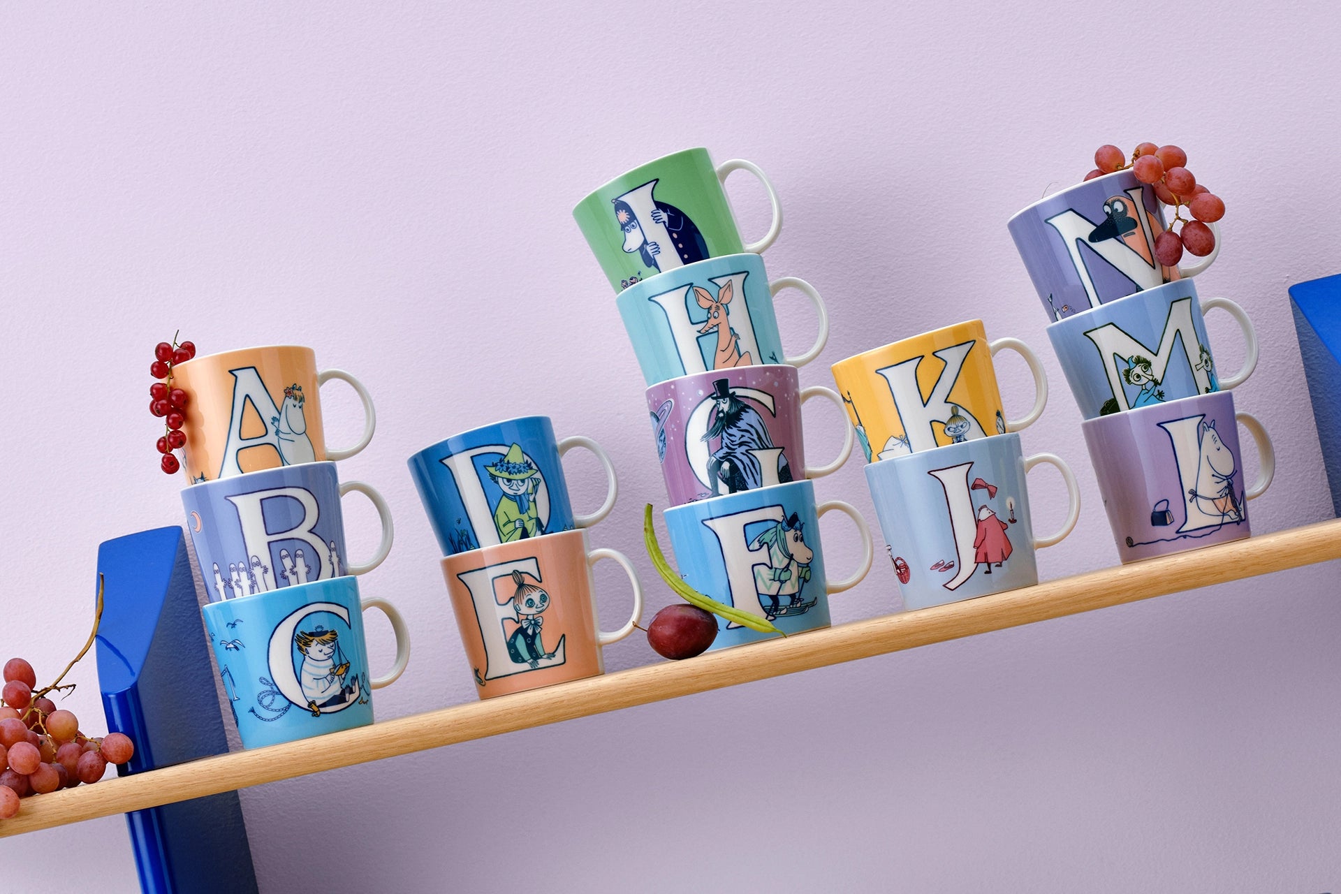 Colorful Moomin Arabia alphabet mugs on a shelf against a light purple background