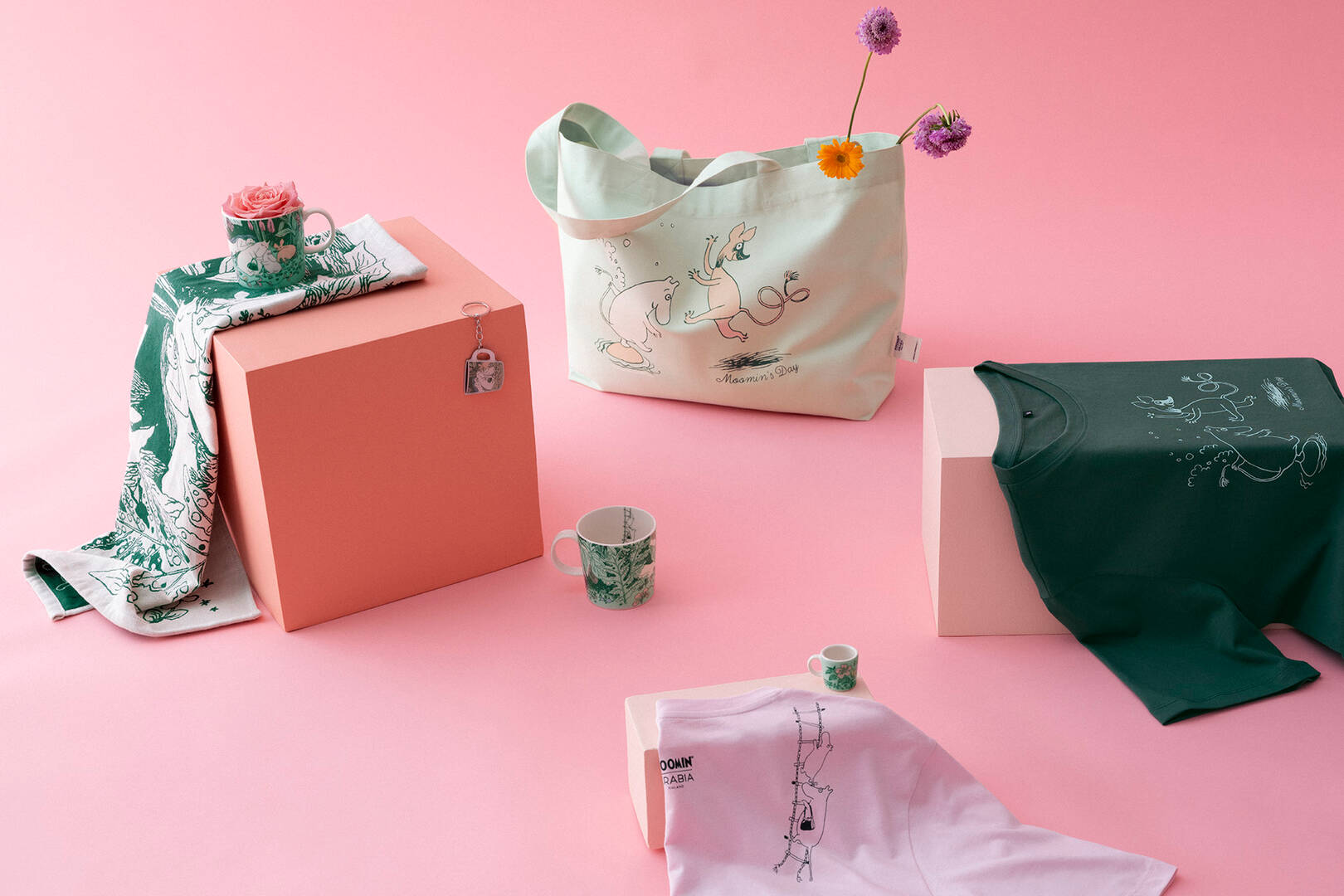 Collection of branded Moomin Day merchandise including a tote bag, t-shirt, and box on a pink background