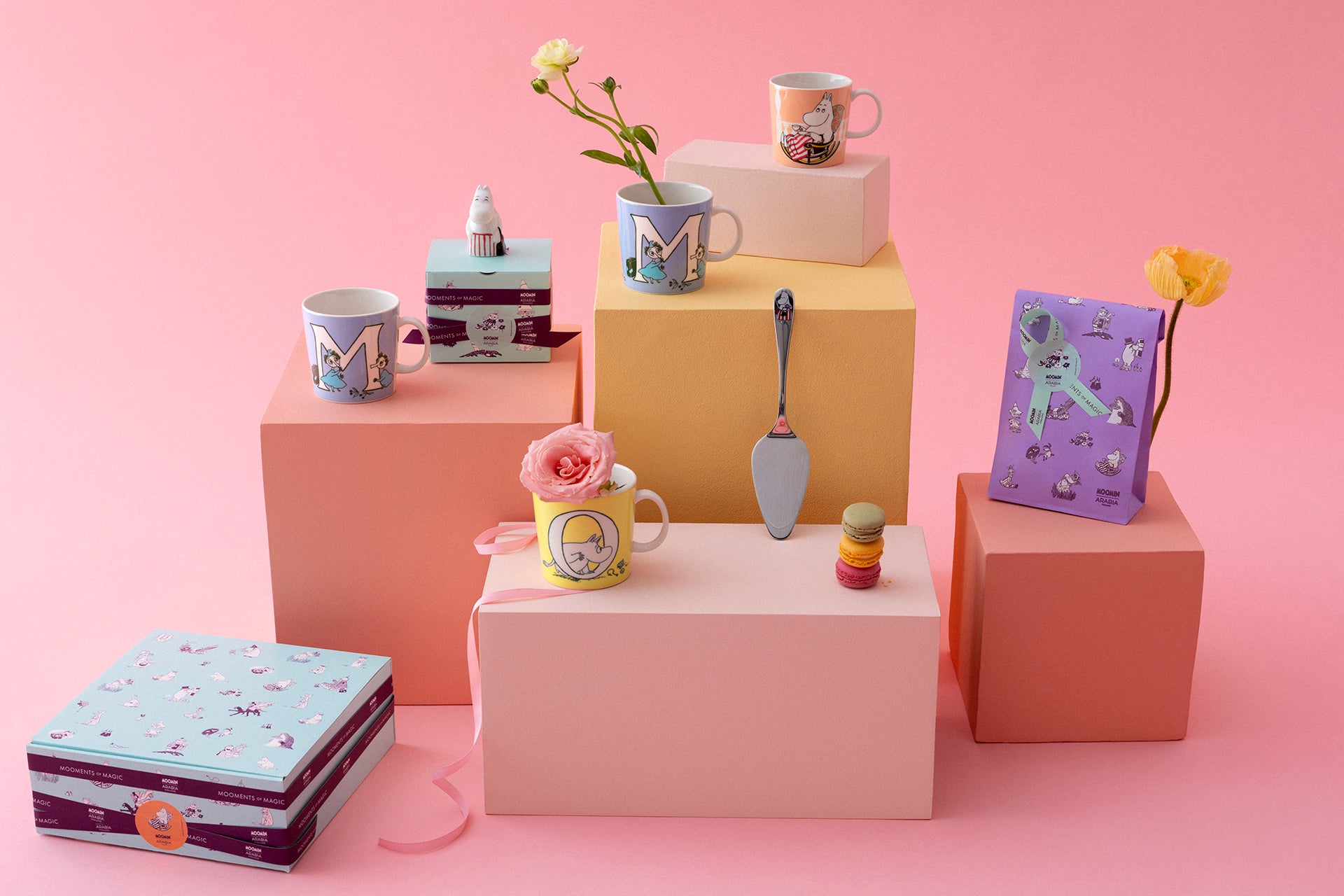Colorful Moomin mugs, gift boxes, and decorative items on pink and beige cubes against a pink background