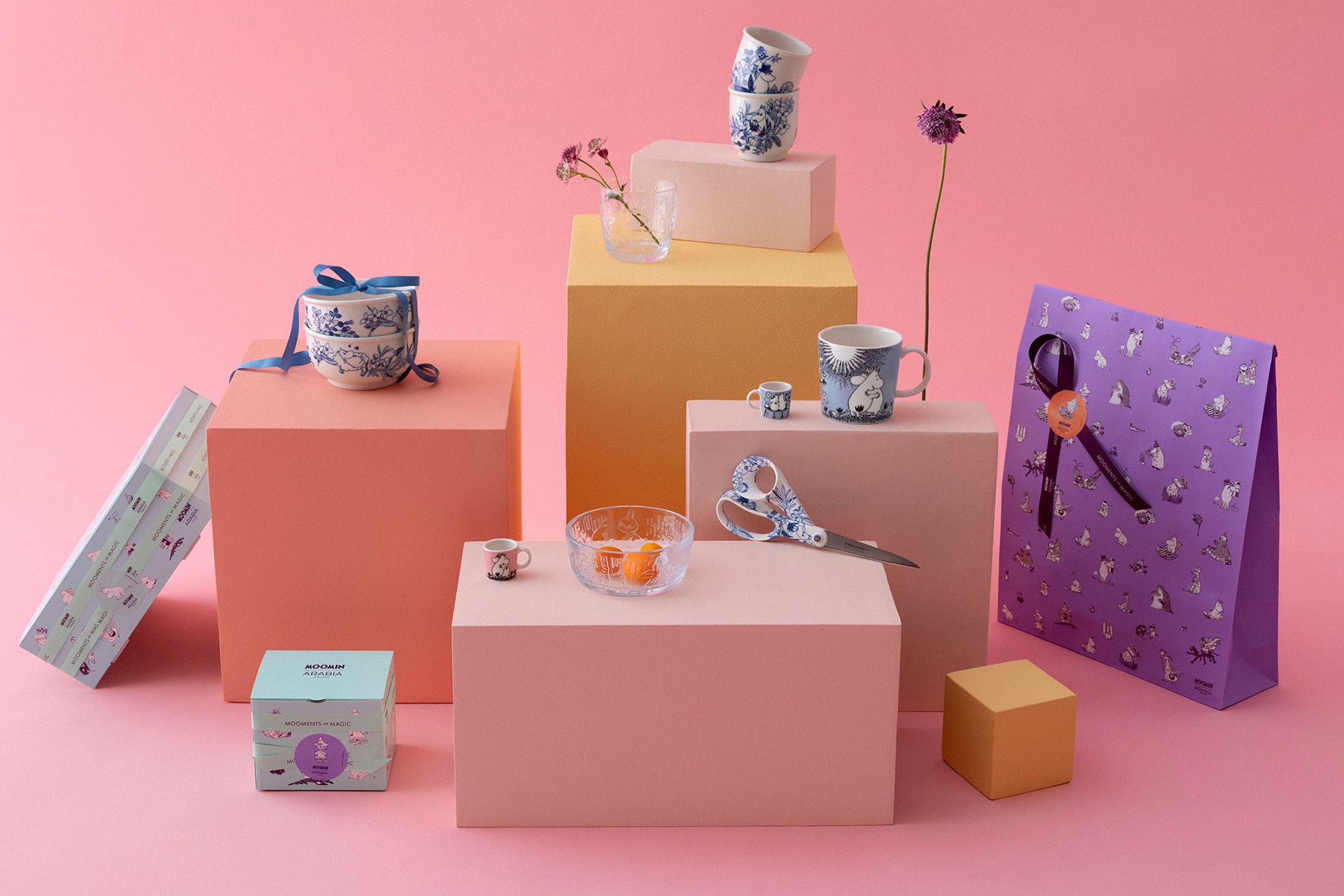 Collection of decorative items including Moomin bowls and cups, a gift bag, and boxes on a pink background