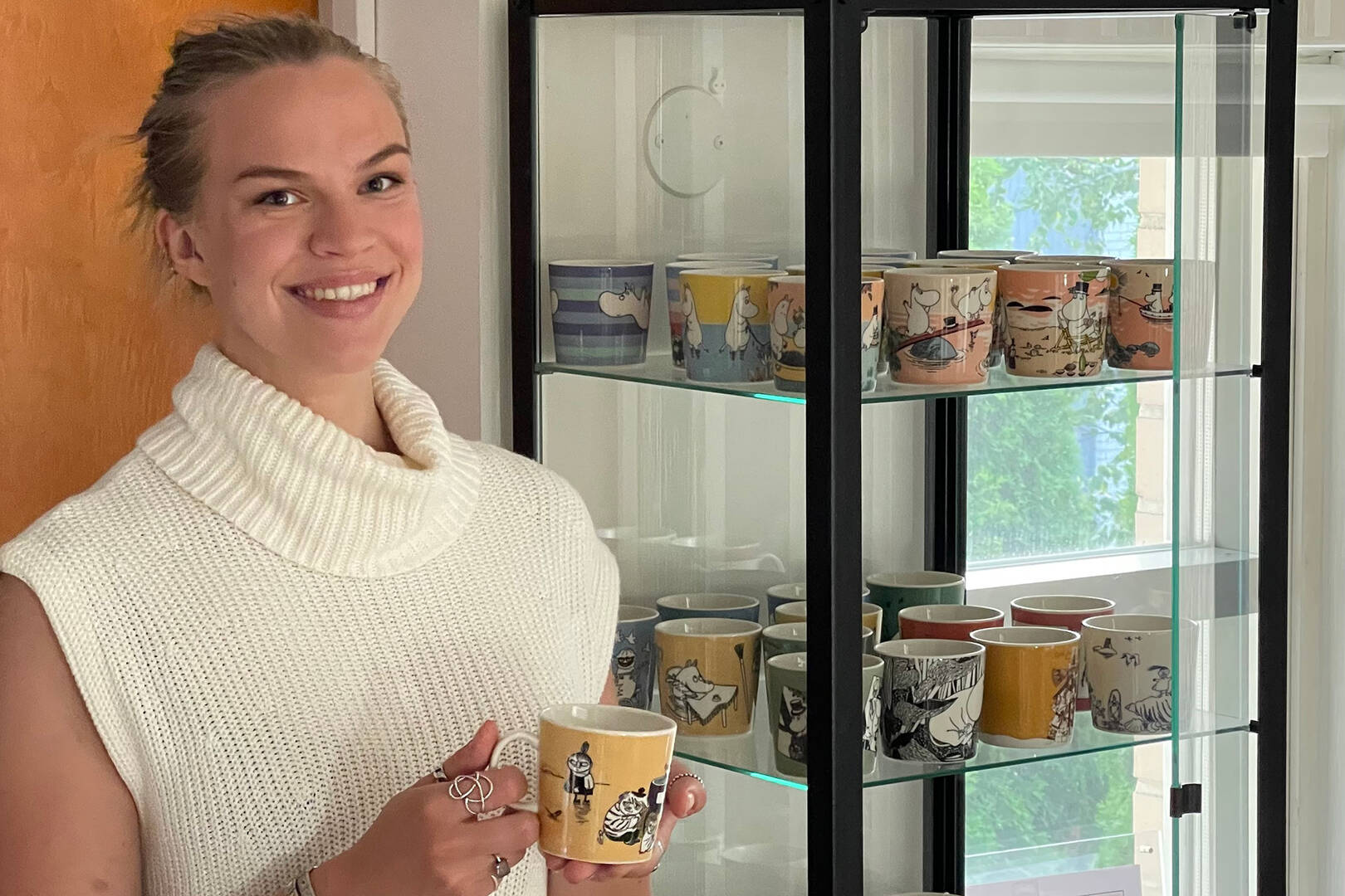 Woman holding a Moomin mug in front of a display cabinet with various mugs.