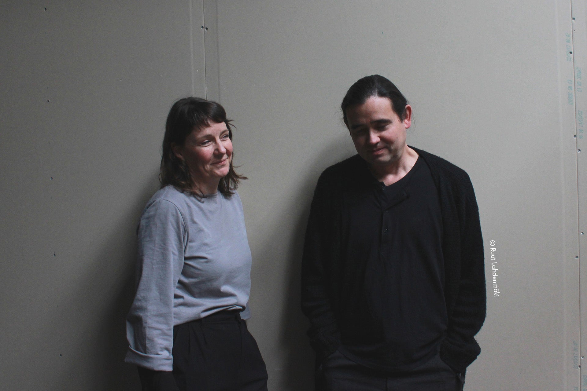 Two people standing against a plain wall.