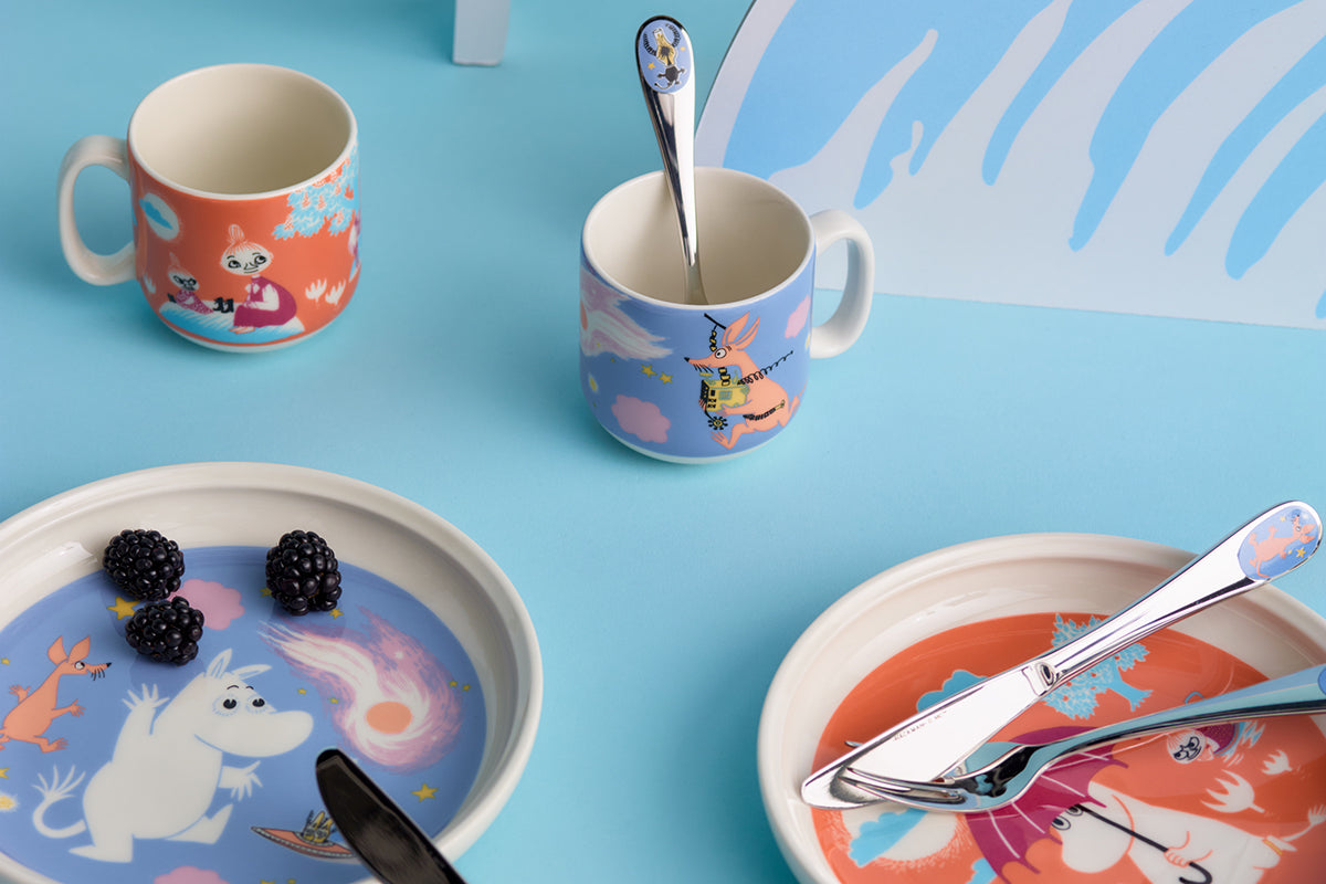 A styled flat-lay of children's Moomin-themed tableware on a light blue surface, including two patterned mugs with spoons, two matching plates with cutlery and blackberries, and a decorative blue-and-white background panel.