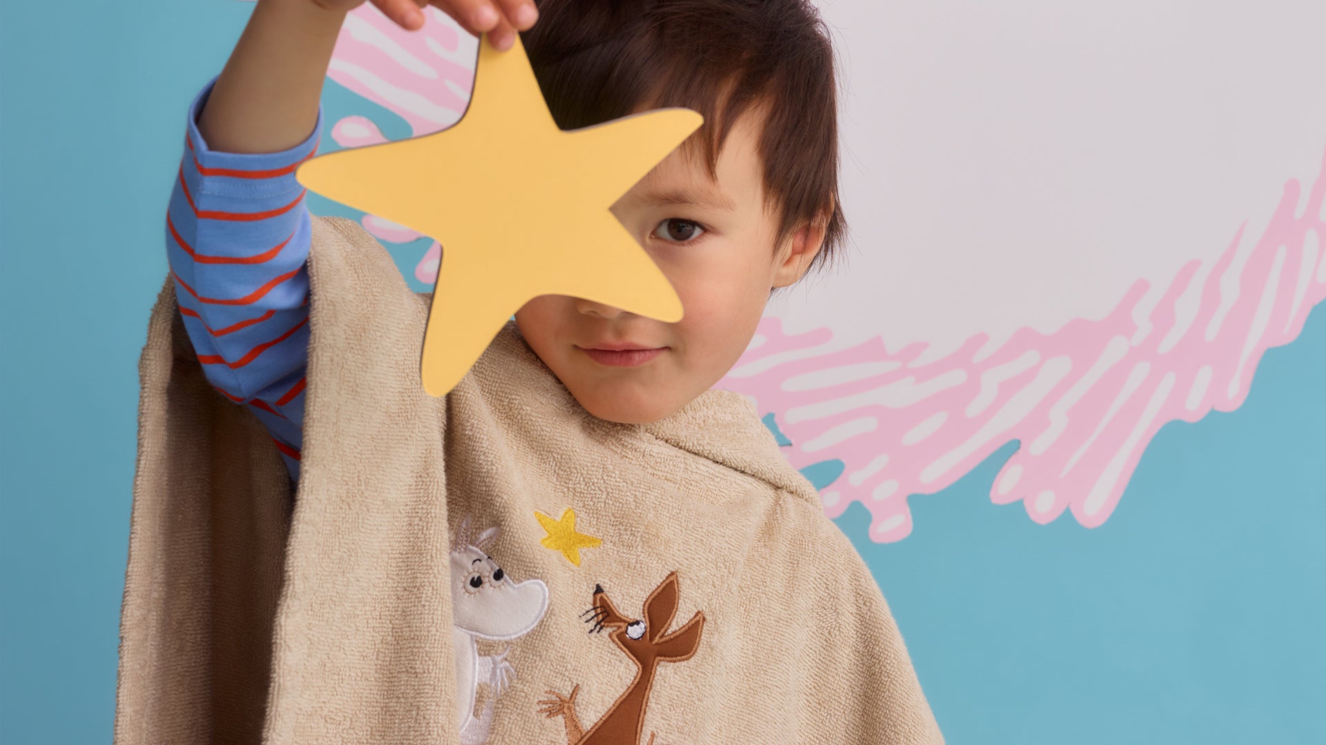 A child wearing a beige Moomin towel cape with cartoon animal designs, holding a yellow star-shaped prop in front of their face, against a blue and pink background.