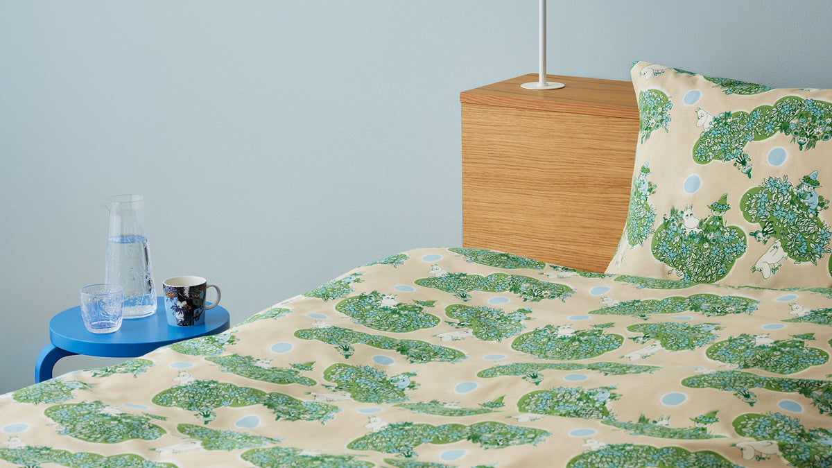 A bed dressed in a beige sheet and pillowcase featuring illustrations of Little My, Moomintroll and Snufkin in a tree, styled in a bedroom setting with a light blue wall, wooden headboard, and a small blue side table with a glass bottle and mug.