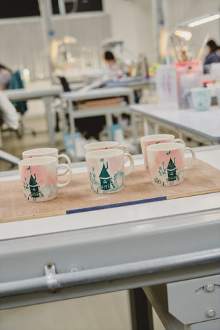 Four Home-mugs with decorative hand-painted designs of Moomin-house on a wooden board in a workshop setting.