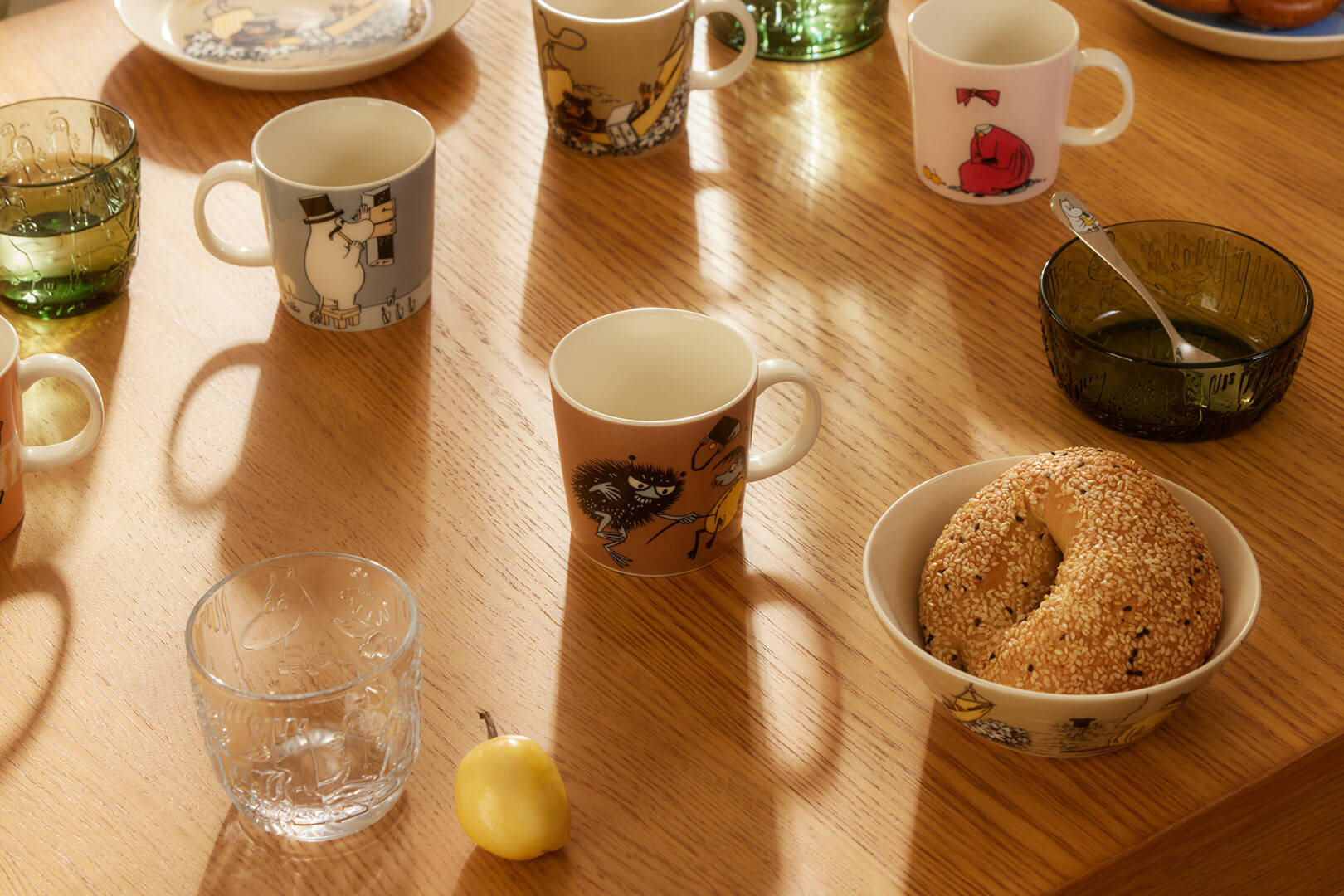 Table with Moomin classics mugs, a bowl of bread, mystical forest tumblers and a pear on a wooden surface