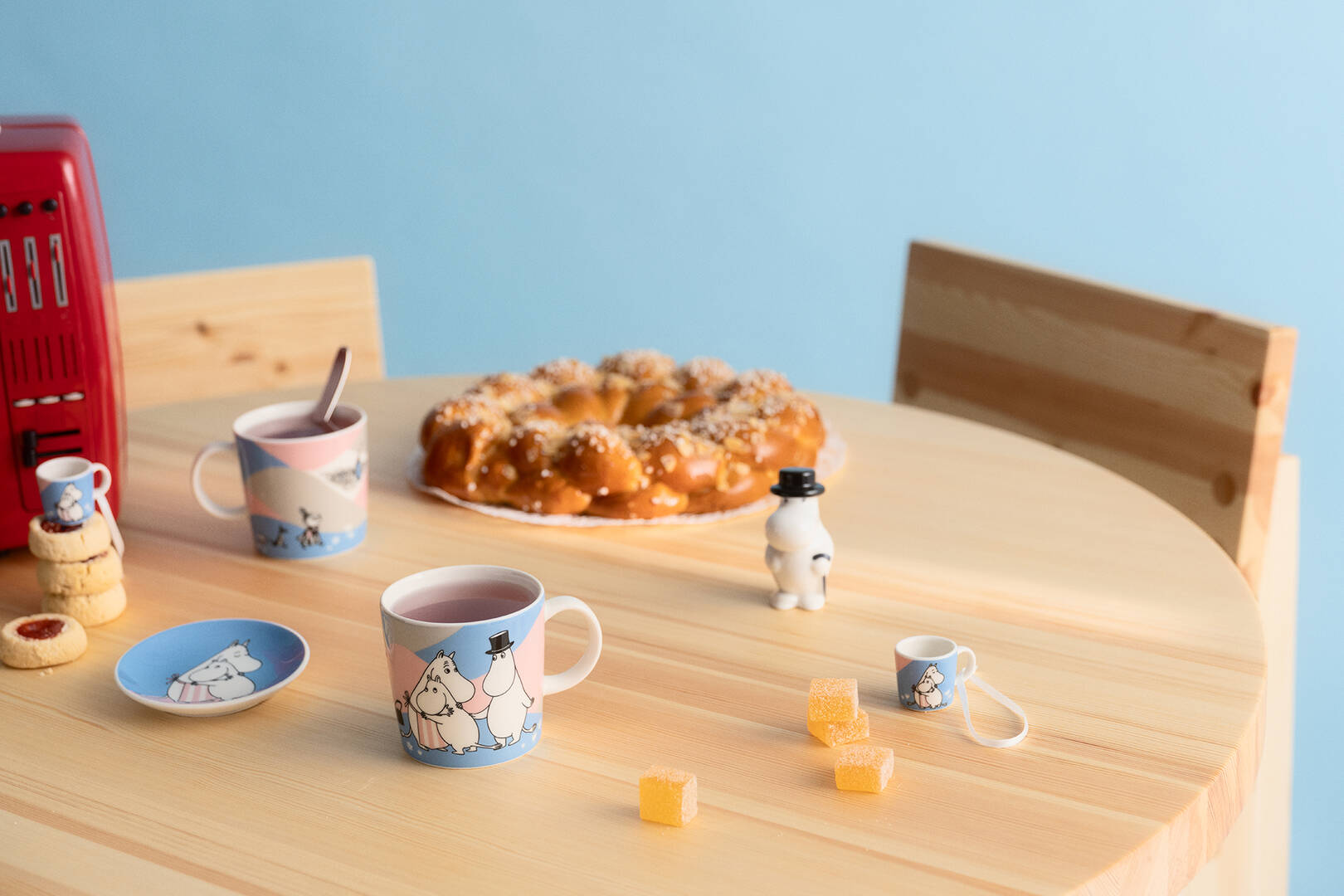 Round wooden table with Hom eat last moomin mugs, a bun crown, and small figures on a light blue background