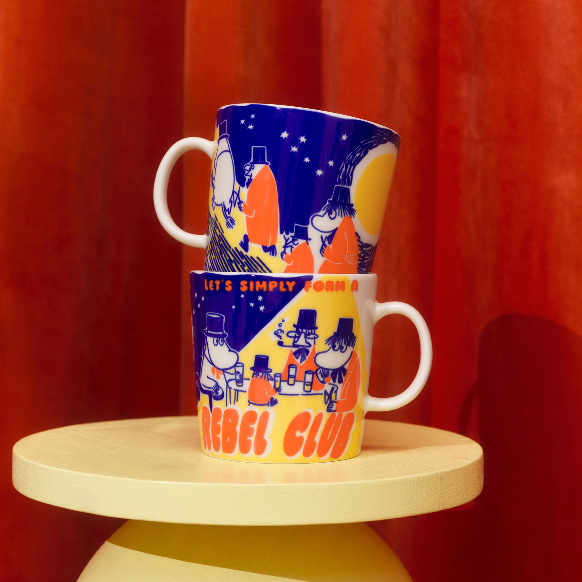 Moomin mug 0,4L Rebel Club - TwoRebel Club mugs stacked on a wooden stool against a red curtain background