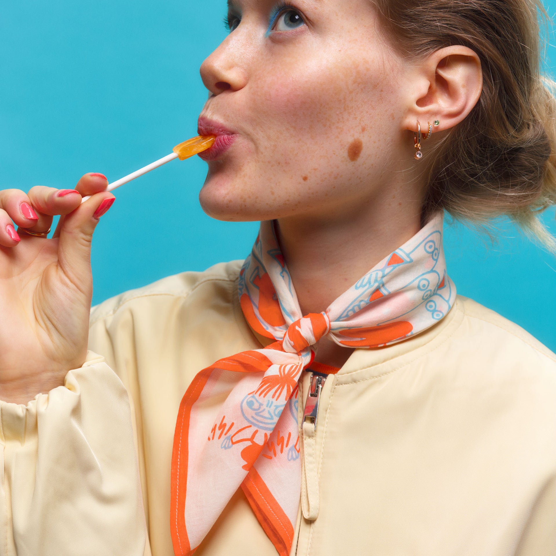 Moomin scarf Party Queue - Woman wearing a Rebel Club scarf, eating a lollipop against a blue background