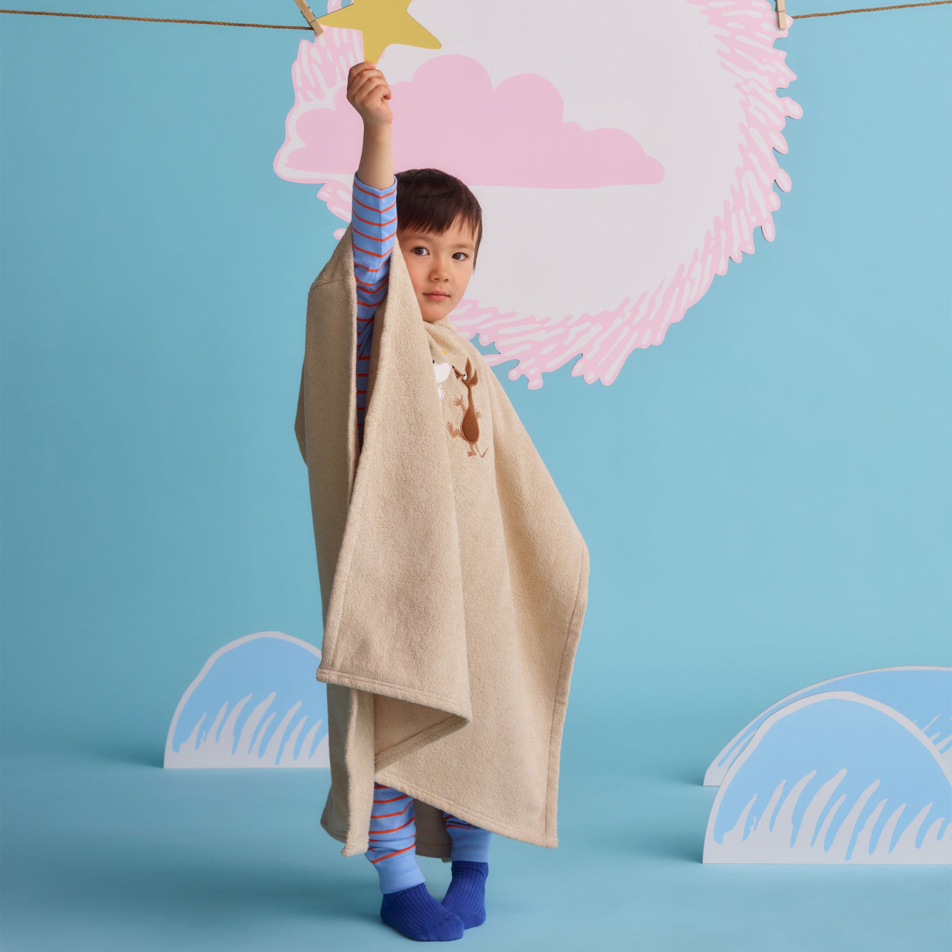Mumin barn badponcho Rymdäventyr - A child wearing a beige poncho towel holding up a star cut out.