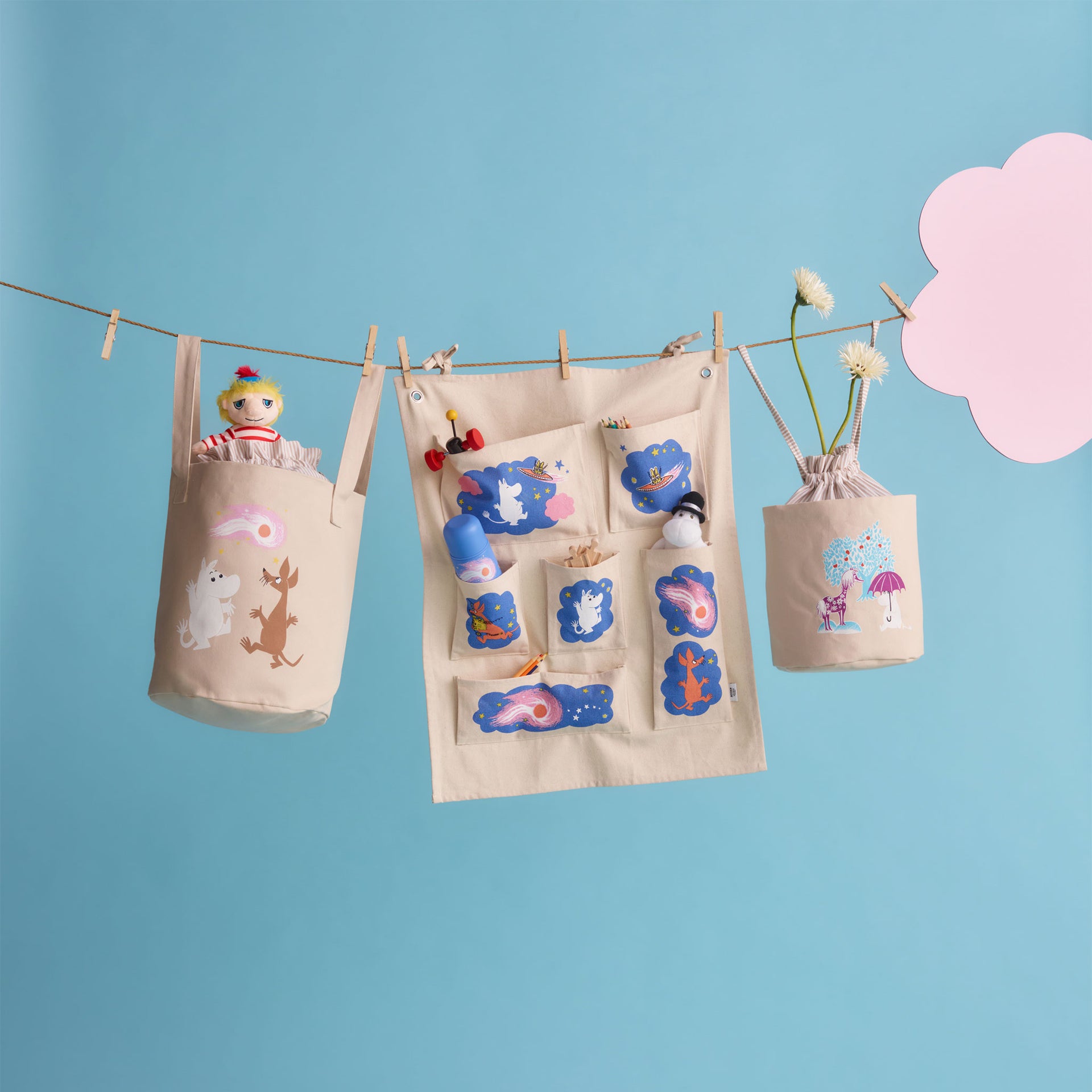 Mumin förvaringskorg 24cm Under paraplyet - A beige fabric storage organizer hanging from a clothesline with wooden clips, decorated with colorful patches and holding plush toys, shown against a light blue background with a pink cloud cutout.