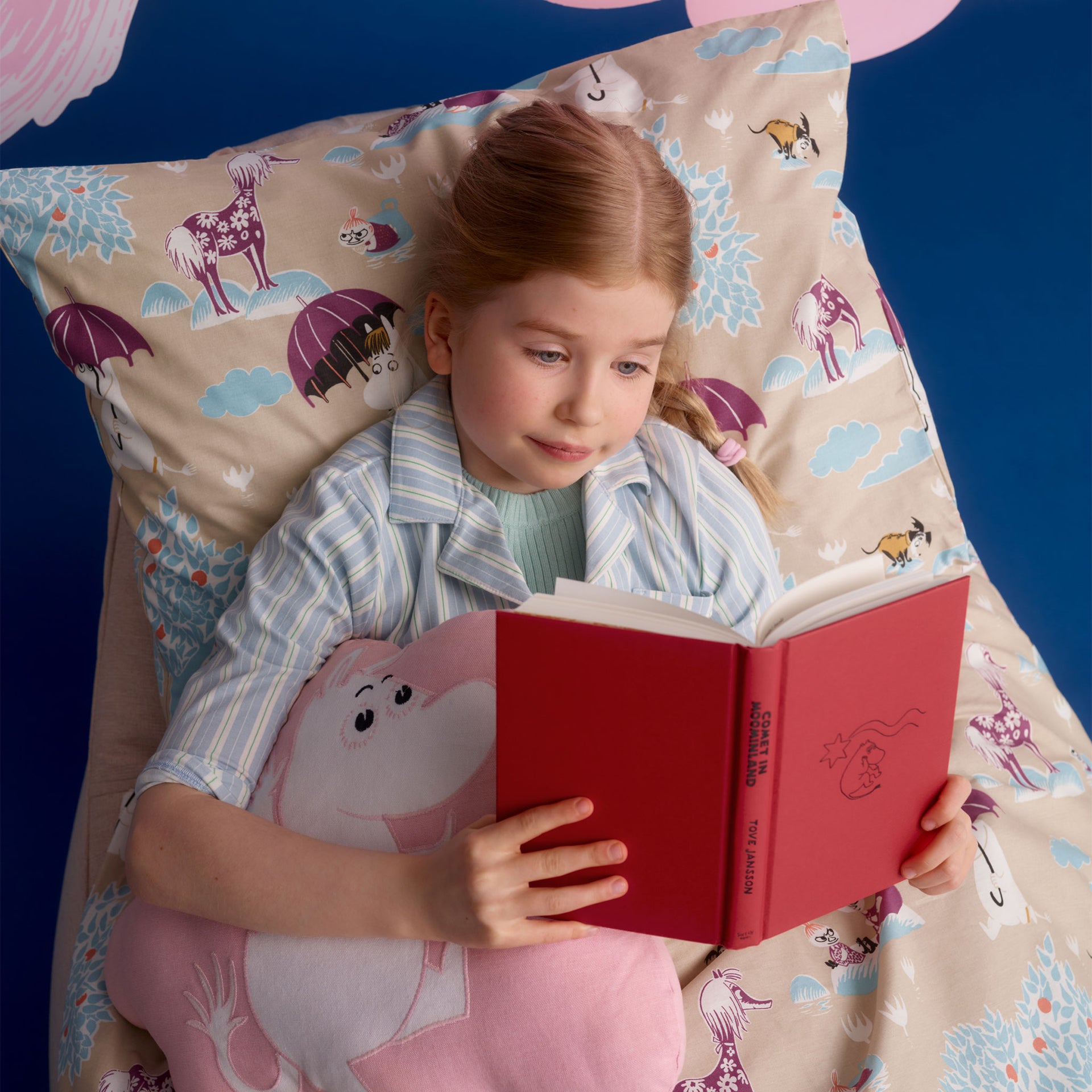 Mumin kudde 37x47cm Rymdäventyr - A young girl with red hair lies in bed reading a red book, surrounded by matching beige bedding with Under the Umbrella illustration, and a pink plush pillow.