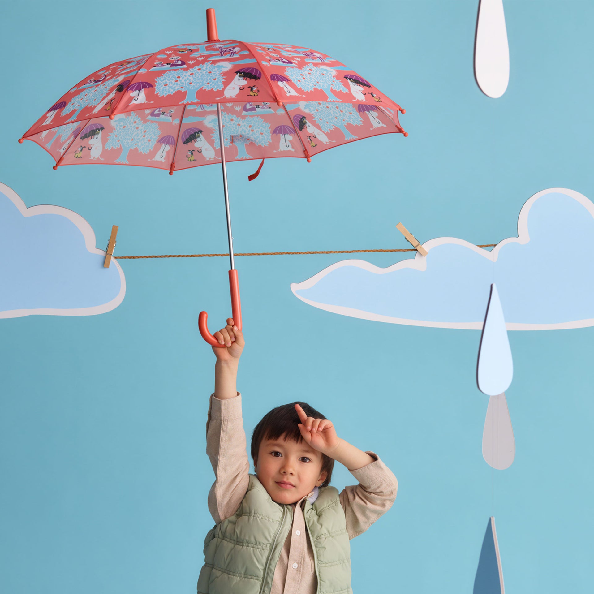 Mumin barn paraply Under paraplyet - A child wearing a light green puffer vest holds up a red umbrella with an Under the Umbrella print, set against a blue backdrop with paper clouds and raindrops.