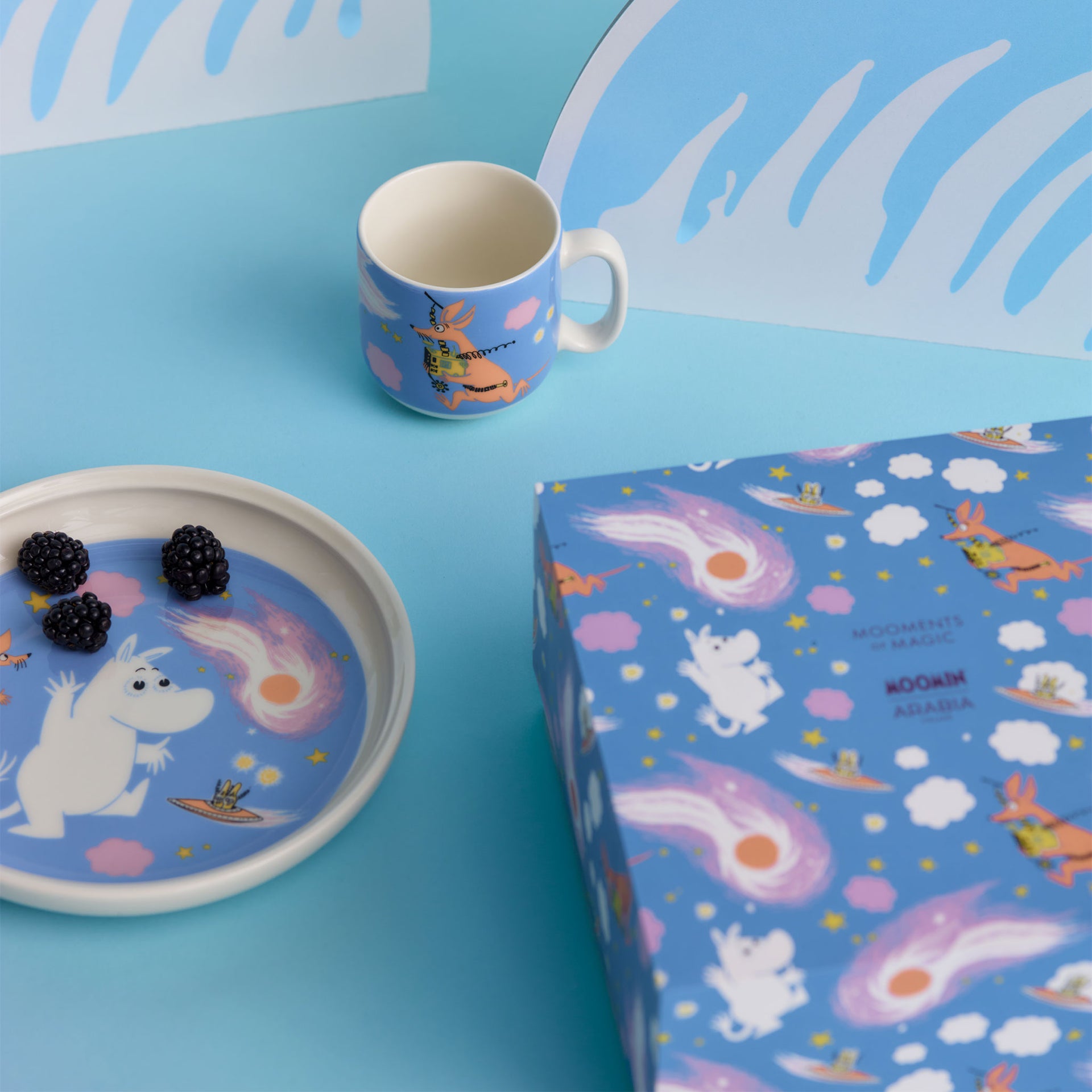 Mumin Barnset Rymdäventyr - A Space Adventure-themed tableware set on a bright blue background, including a mug, a plate with blackberries, and a matching gift box. 