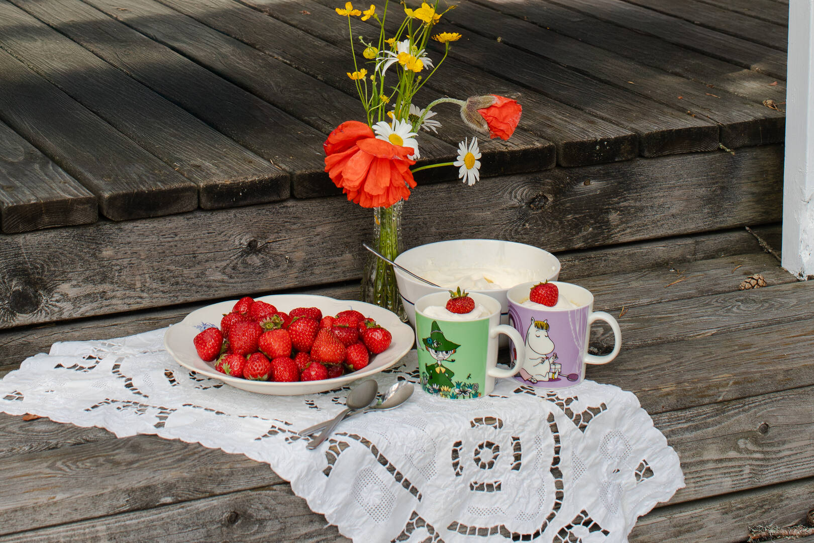 Strawberries on a plate with two Moomin mugs and a vase of flowers on a wooden surface