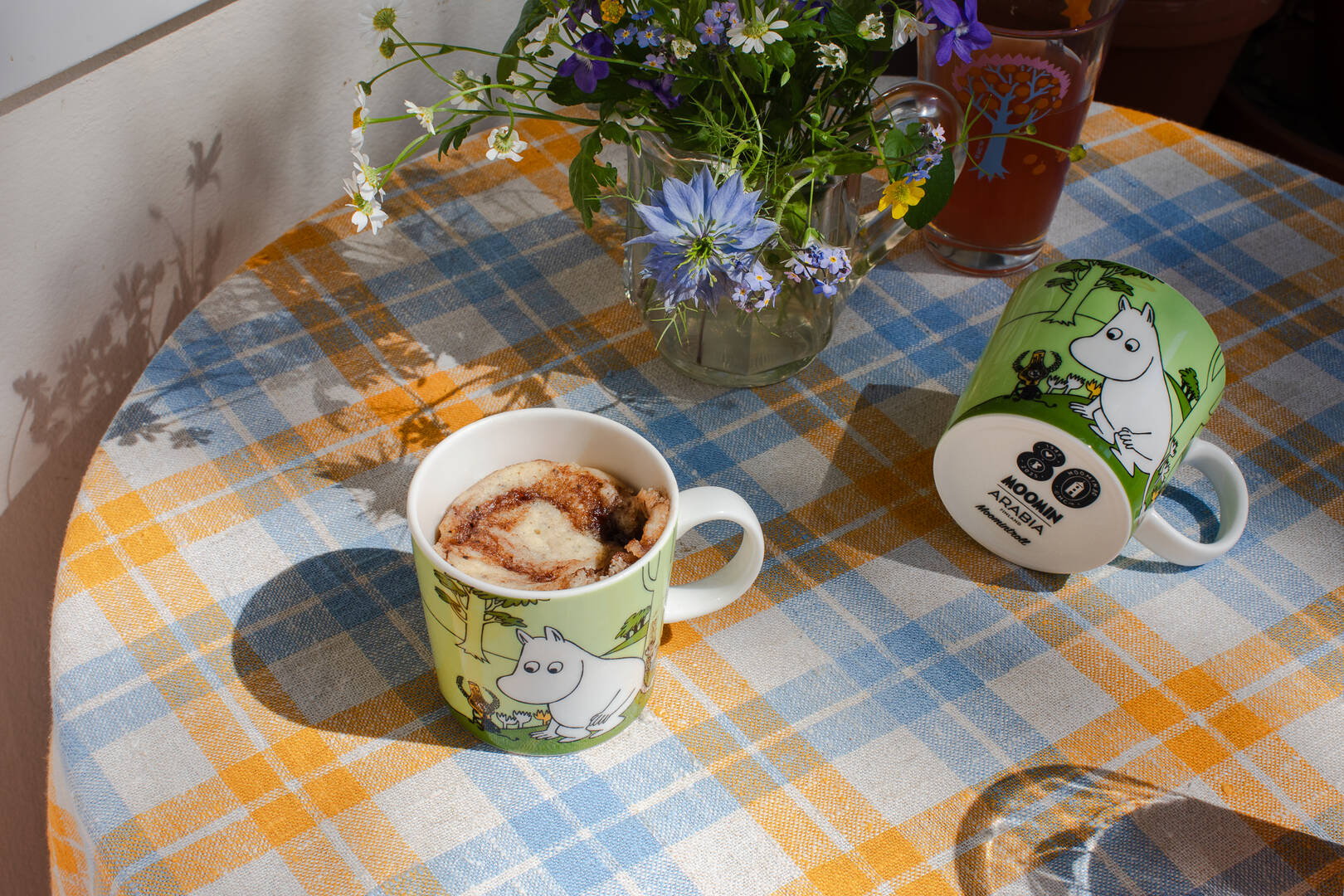 Two Moomintroll-themed mugs on a checkered tablecloth with a vase of flowers and a glass of juice.