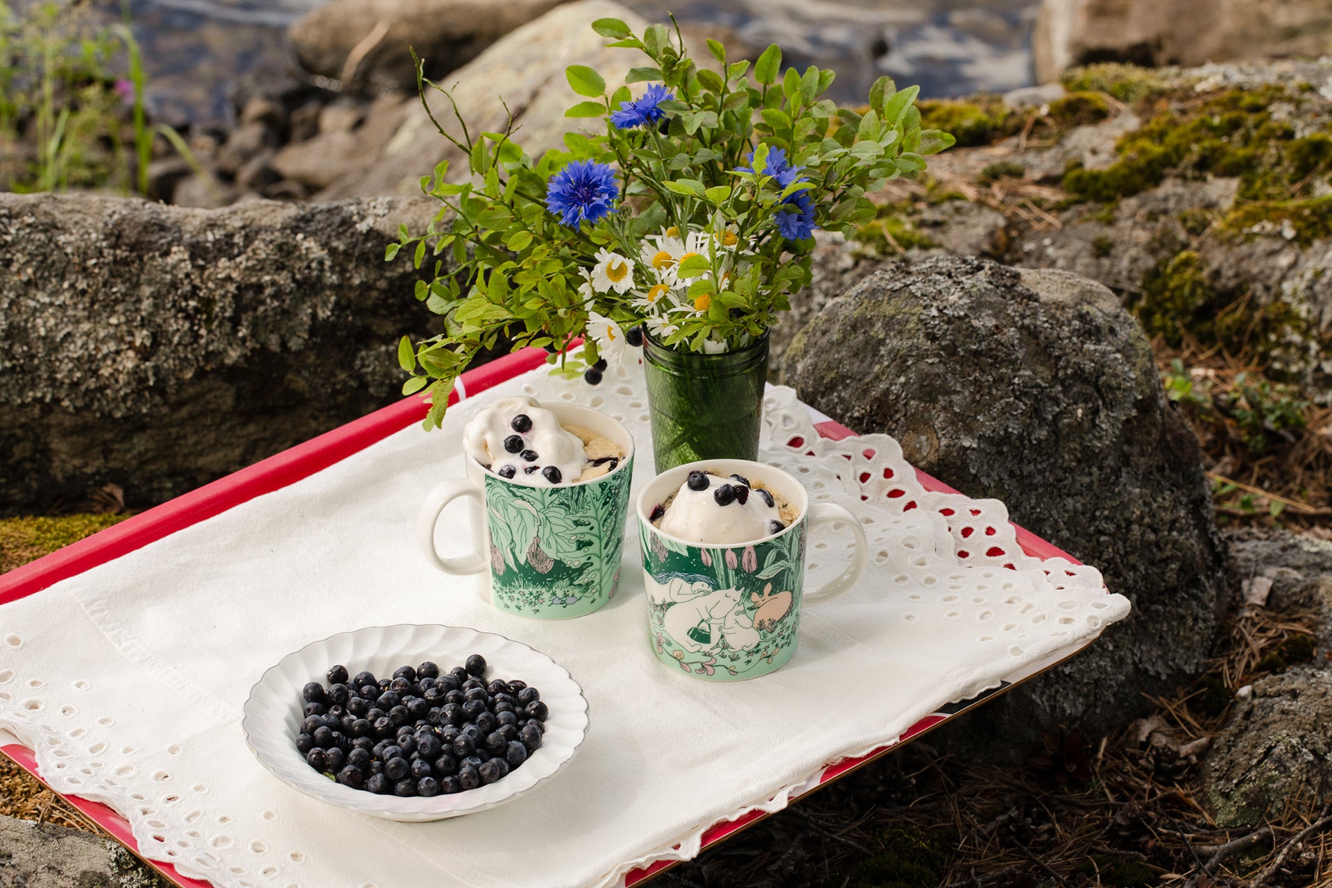 Cups with Moomin Day forest designs, a bowl of bilberries, and a vase of flowers on a white cloth outdoors.