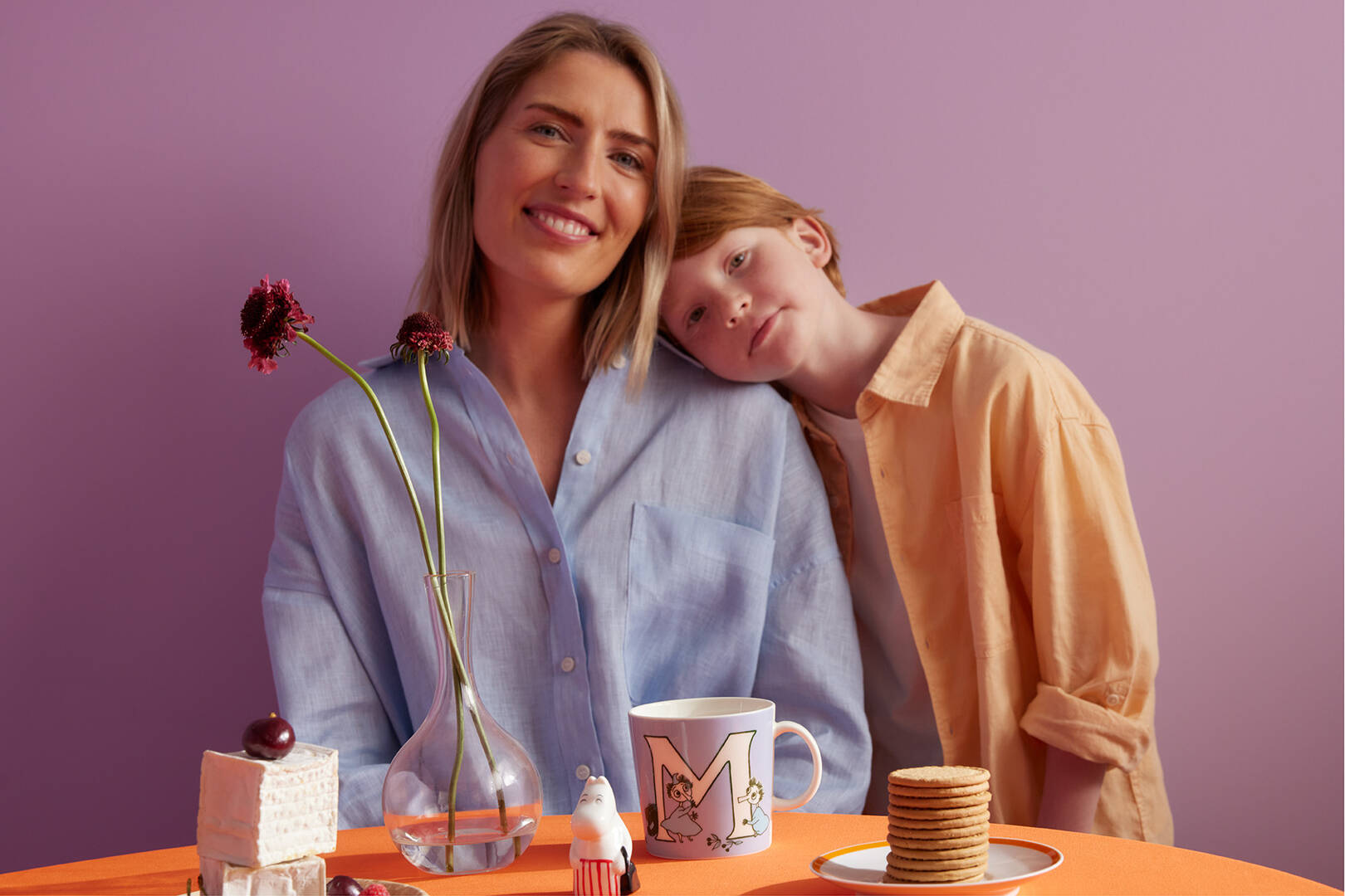 Two people, a mother and a son leaning on a table with a Moomin abc M-mug, cookies, and a vase against a purple background