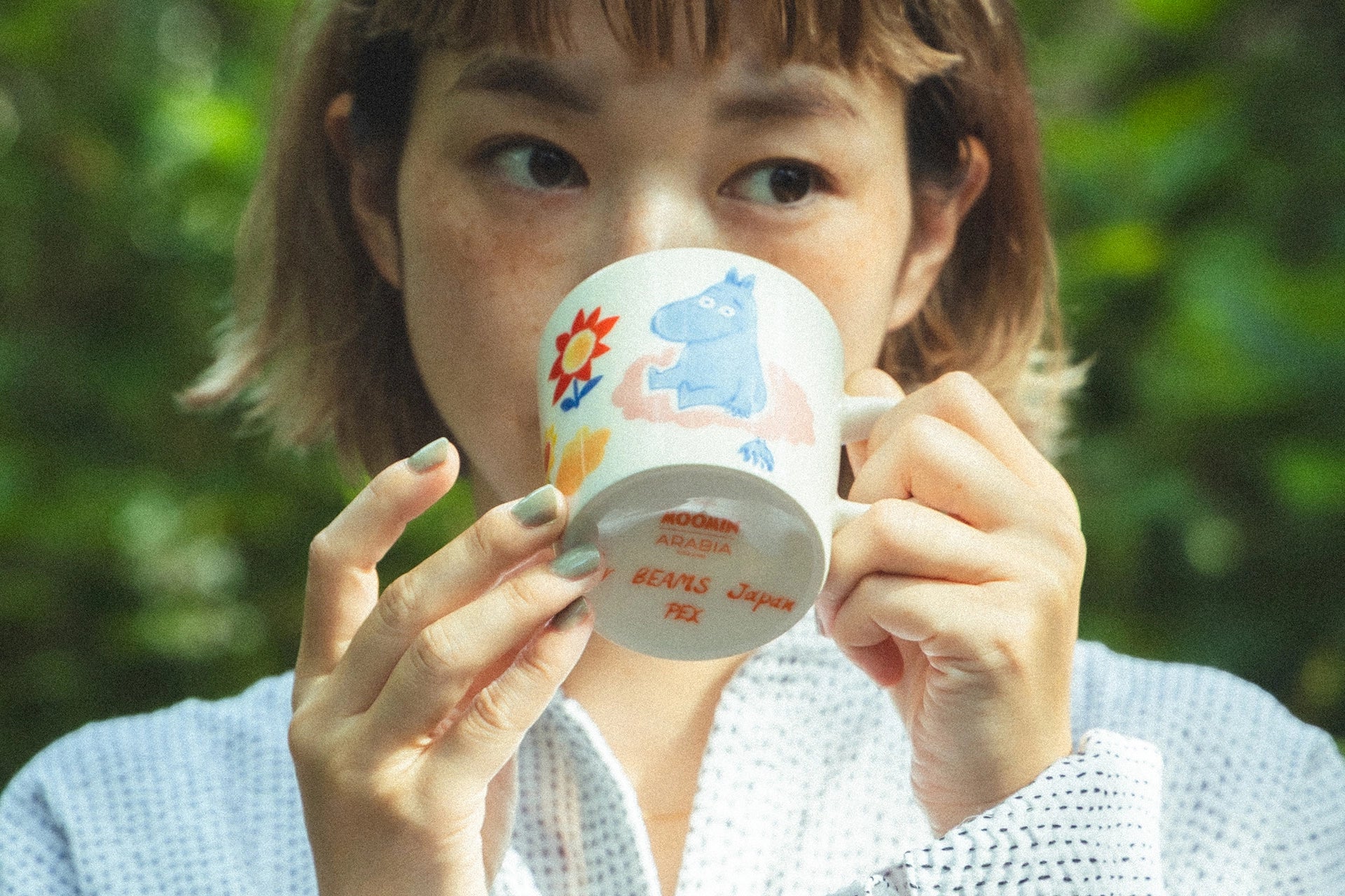 Person holding a colorful beams x moomin arabia mug with a moomintroll design against a blurred green background