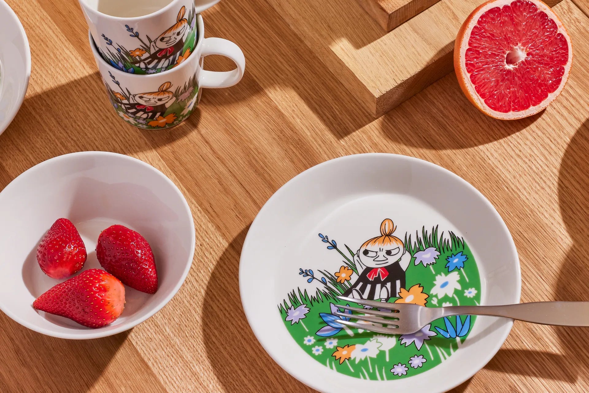 Moomin Arabia plate with Little My on a meadow design on a wooden table with strawberries and a half-slice of grapefruit.