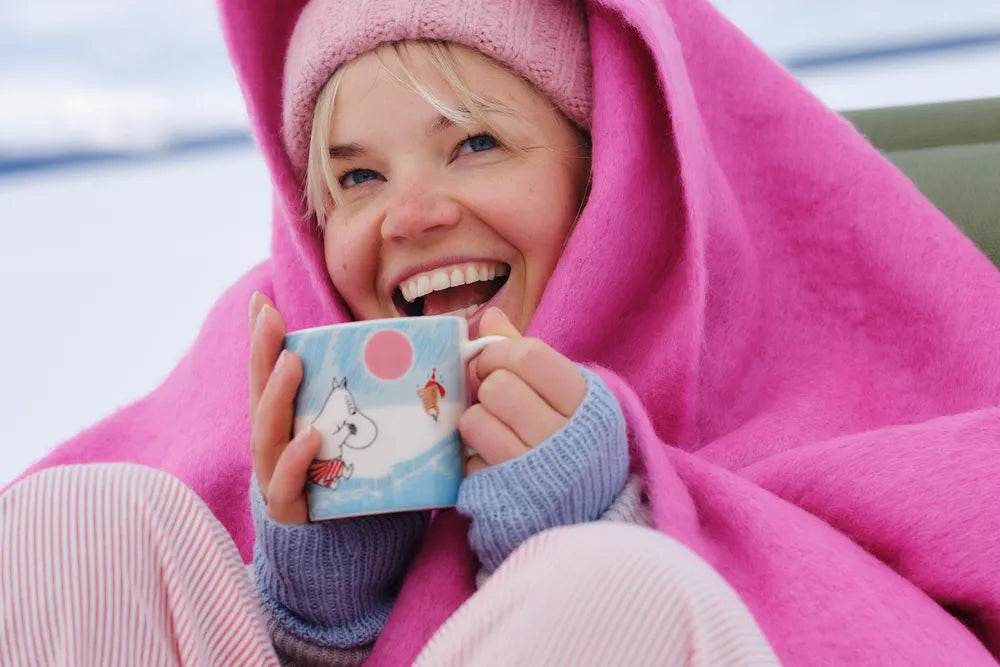Person wrapped in a pink blanket holding an ice swimming mug with a moomintroll design, outdoors.