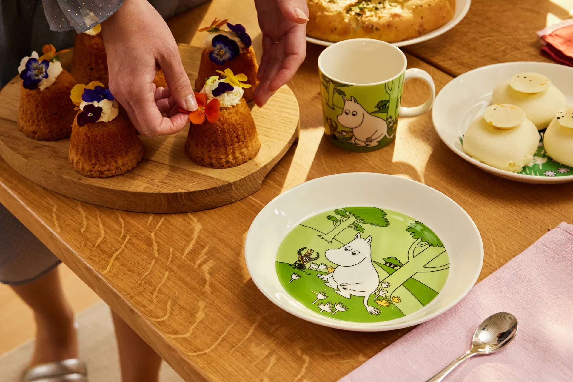 Table setting with small cakes, a mug, and a plate with green Moomintroll design on a wooden table.