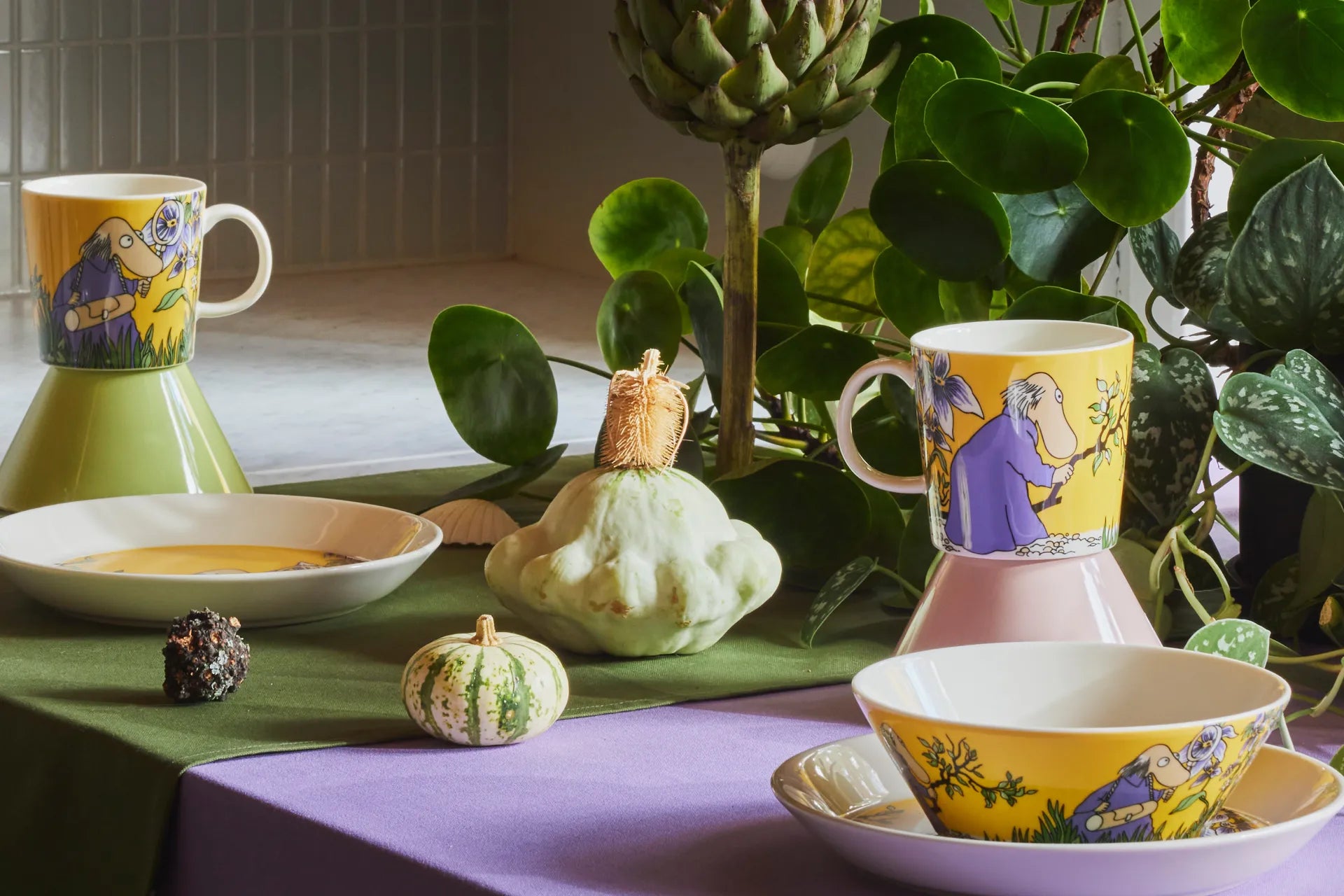 Colorful mugs and bowls with yellow Hemulen design on a table with plants in the background