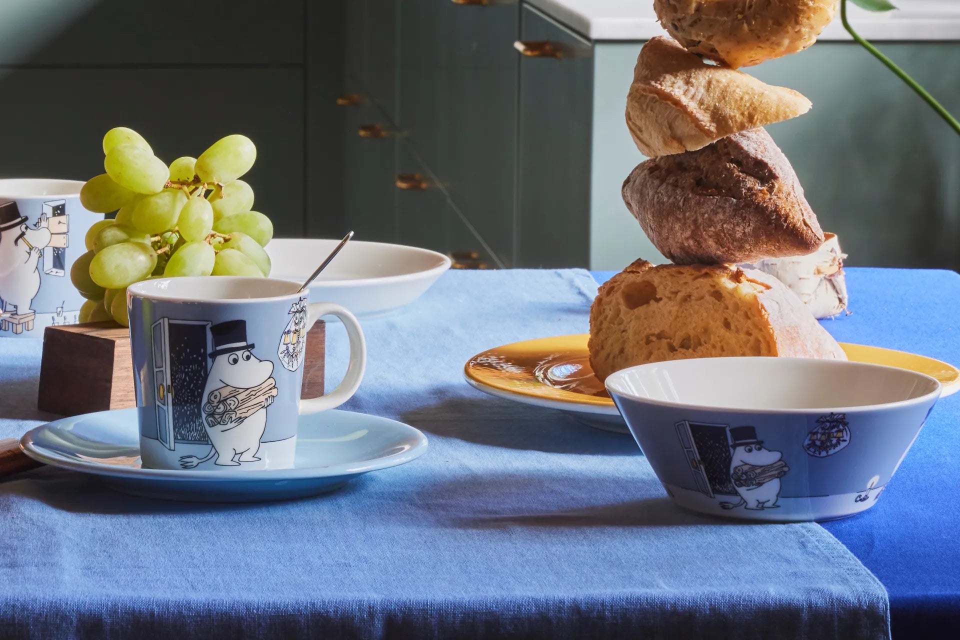 Breakfast scene with a Moominpappa mug, saucer, and bread tower on a blue tablecloth.