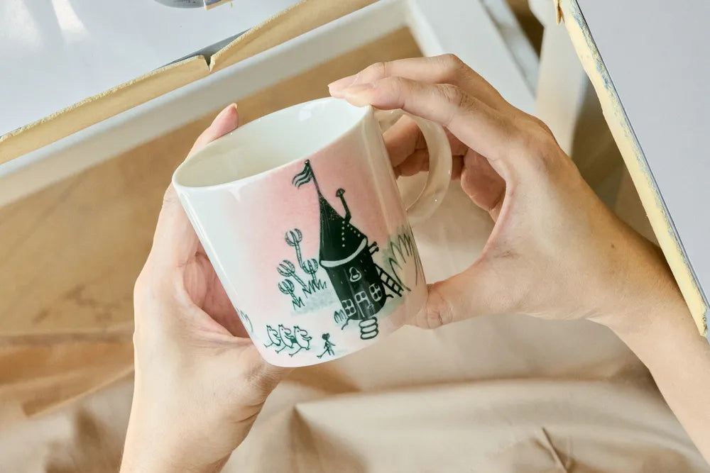 Person holding a mug with a design of a Moomin house and cactus on a wooden surface.
