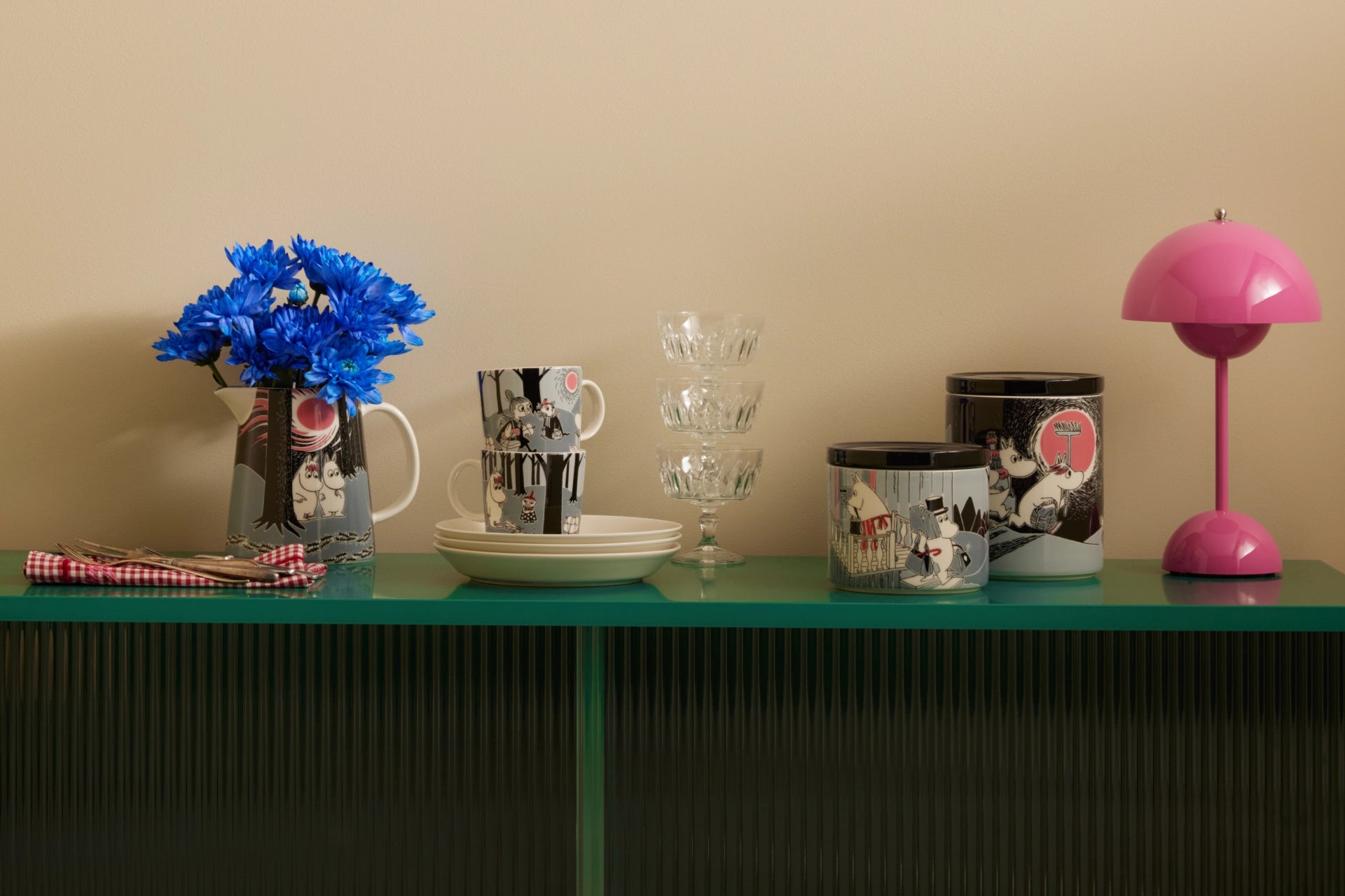 Moomin adventure move items, glasses and a pink lamp on a green table against a beige wall