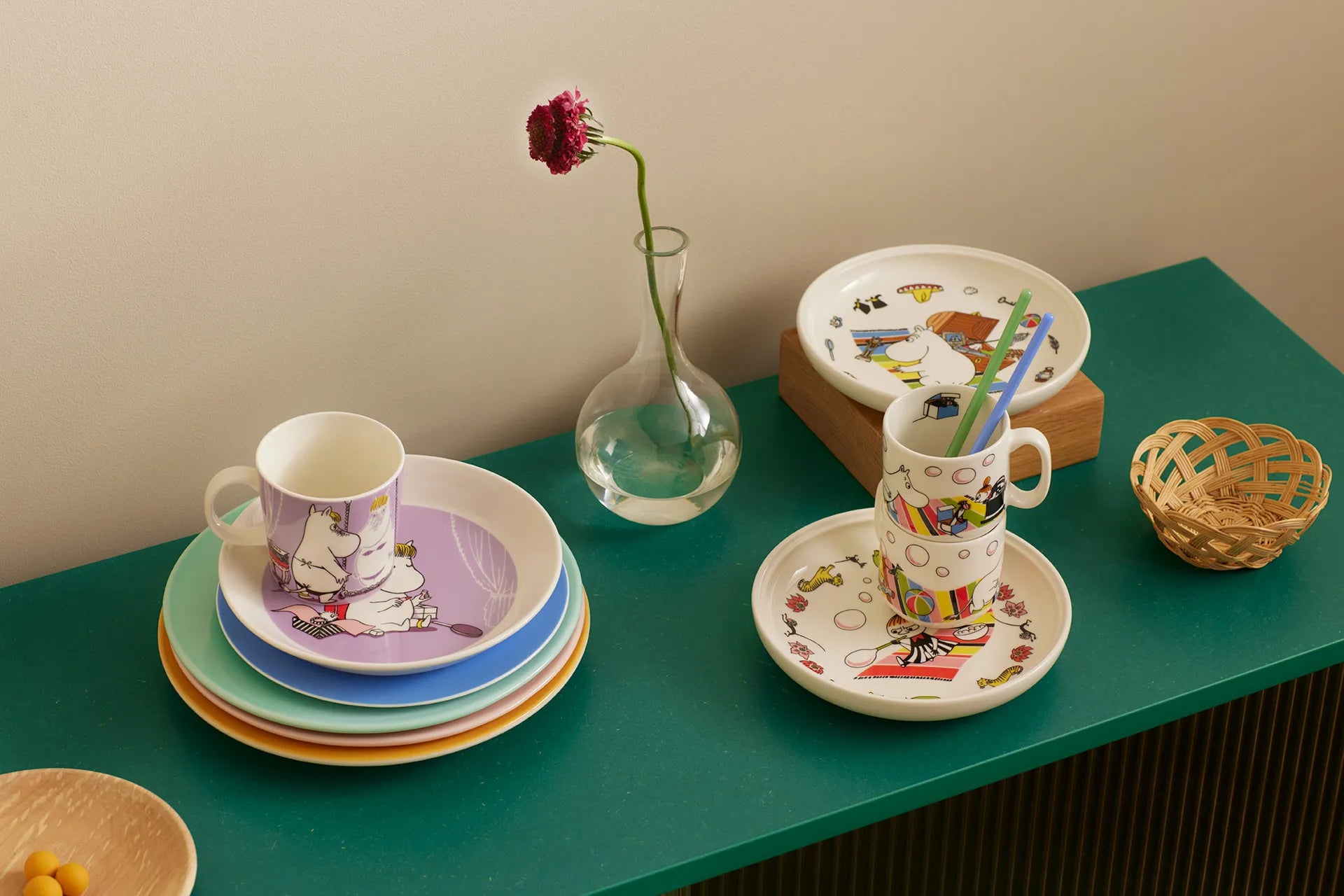 Colorful Moomin children's tableware set on a green surface with a vase and basket.