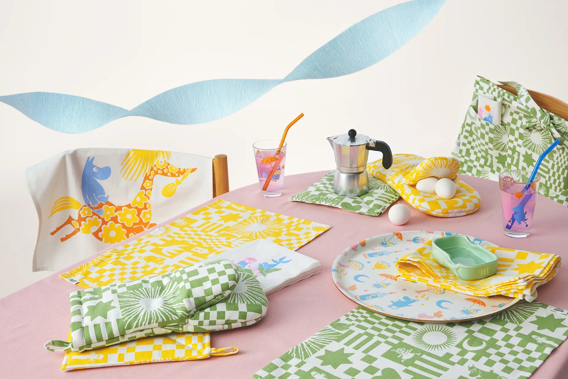 Colorful table setting with Moomin Arabia Party-patterned tablecloth, plates, cups, and napkins on a pink surface.