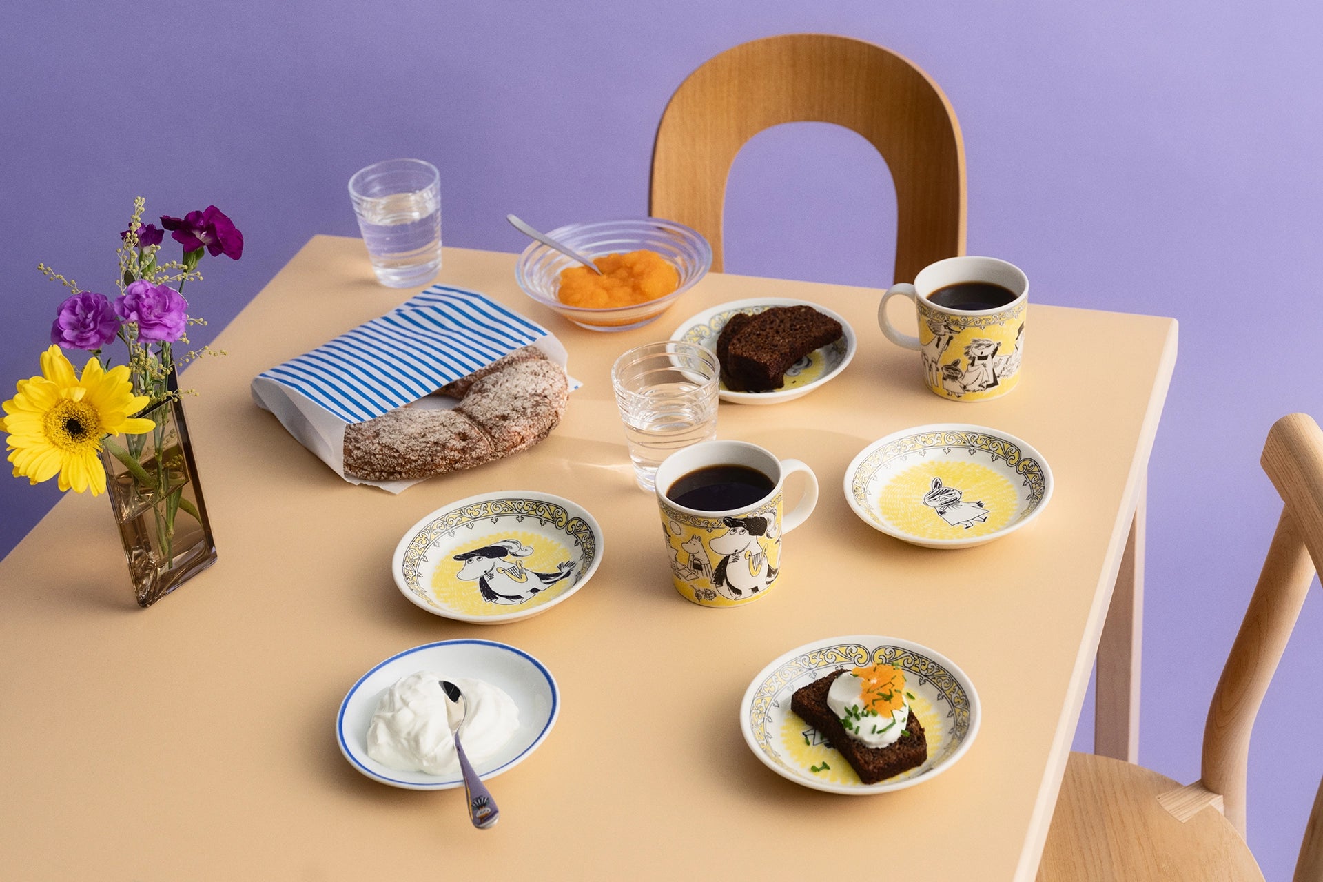 Table setting with various Moomin Scope opera mugs, plates, dishes and a loaf of bread on a purple background