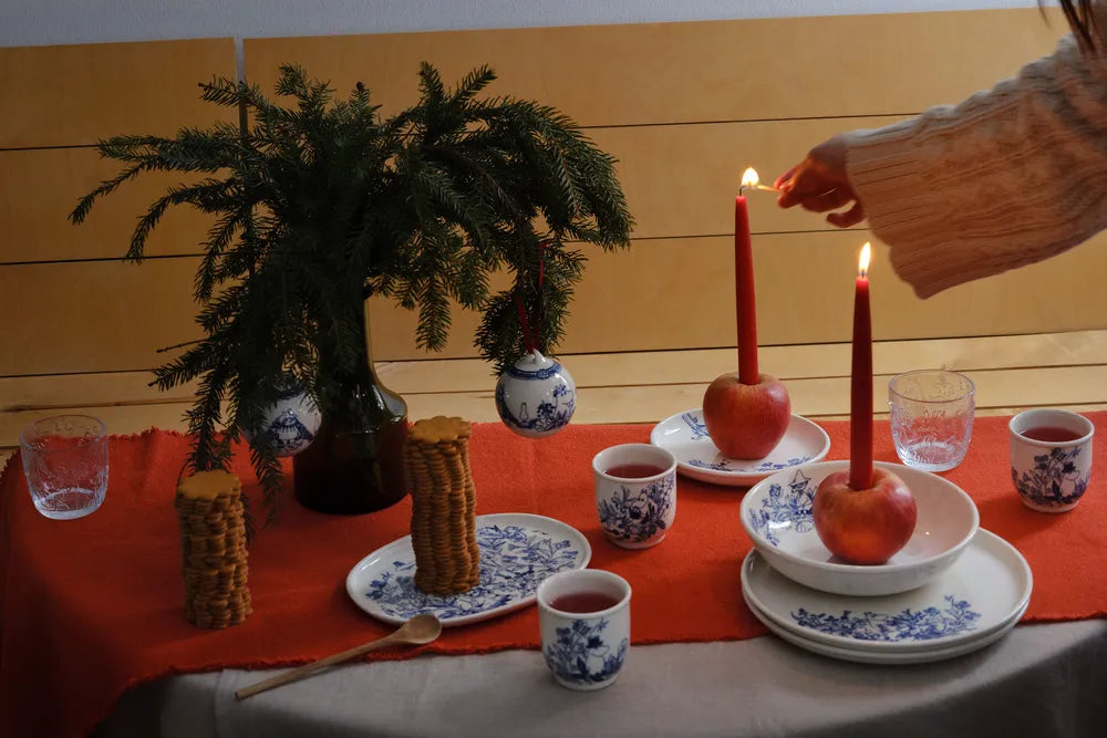 Christmas table setting with red candles, apples, cinnamon cookies and Haru dishes against a wooden wall.
