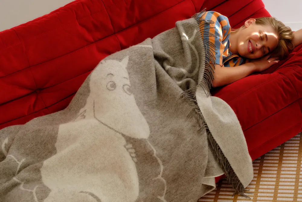 Person lying on a red couch under a beige Moomintroll blanket